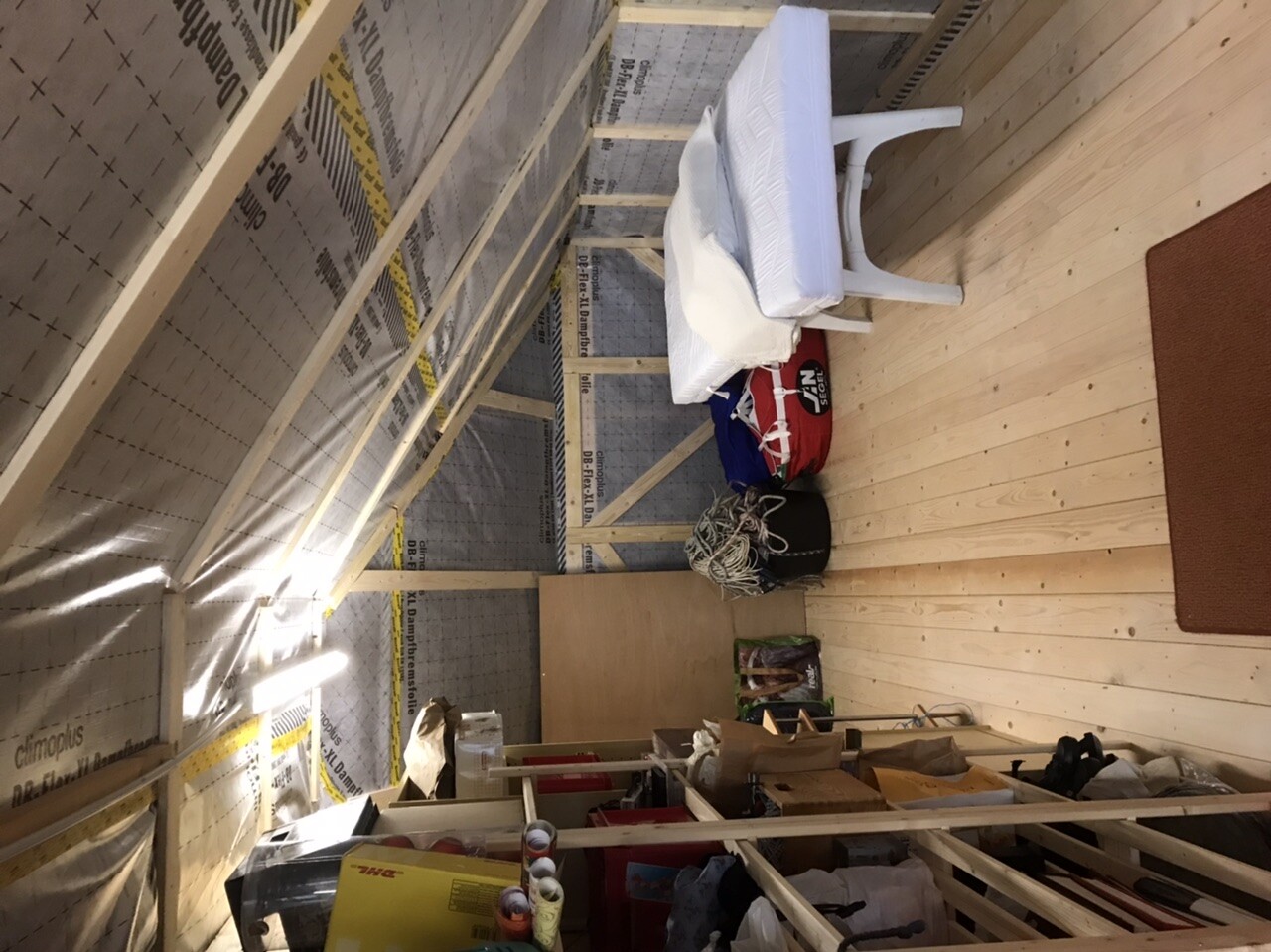 Dachboden mit Holzrahmenkonstruktion, isolierten Wänden, weiße Matratze auf Stuhl, Taschen, Seile.