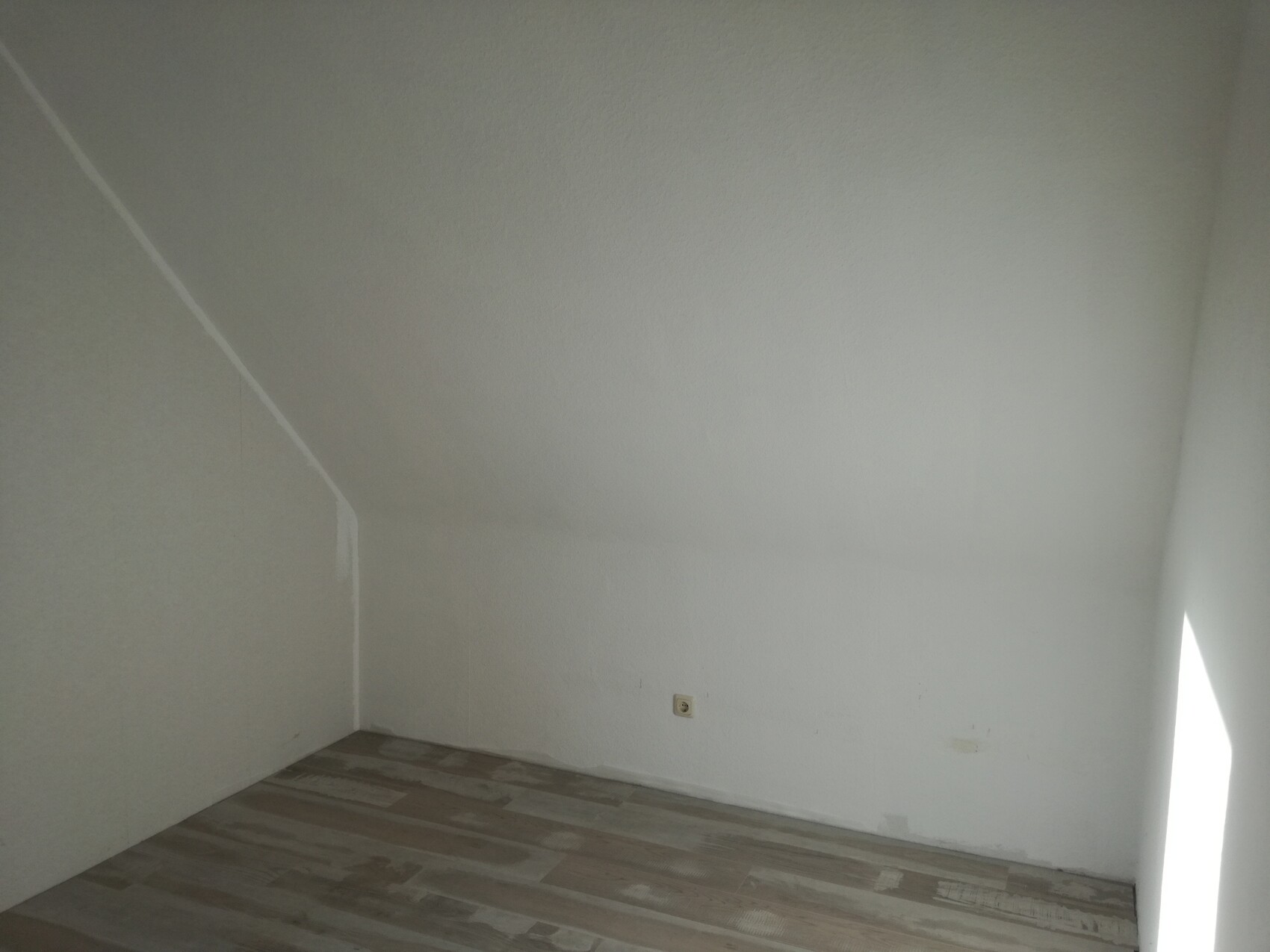 Leerer Raum mit schräger Decke links, helles Licht, Laminatboden, Wandsteckdose.