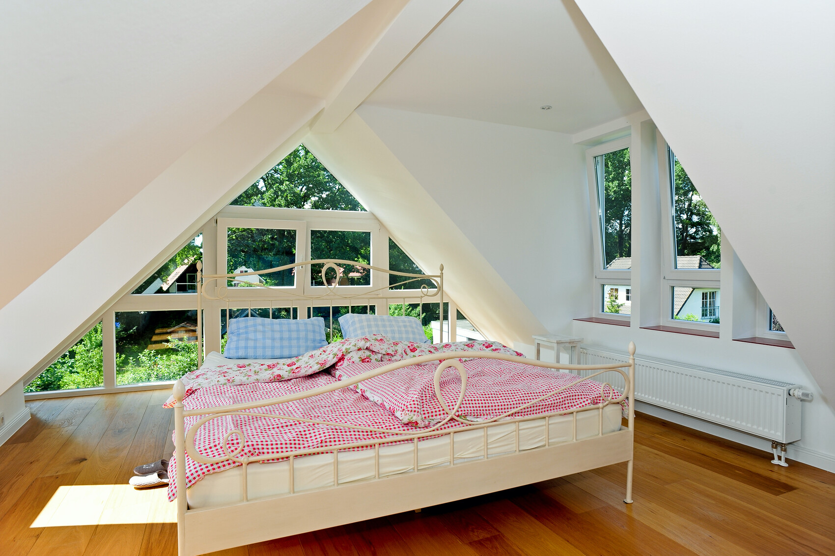 Helles Dachgeschosszimmer mit weißem Metallbett, rosa Bettwäsche und Fenster mit Blick ins Grüne.