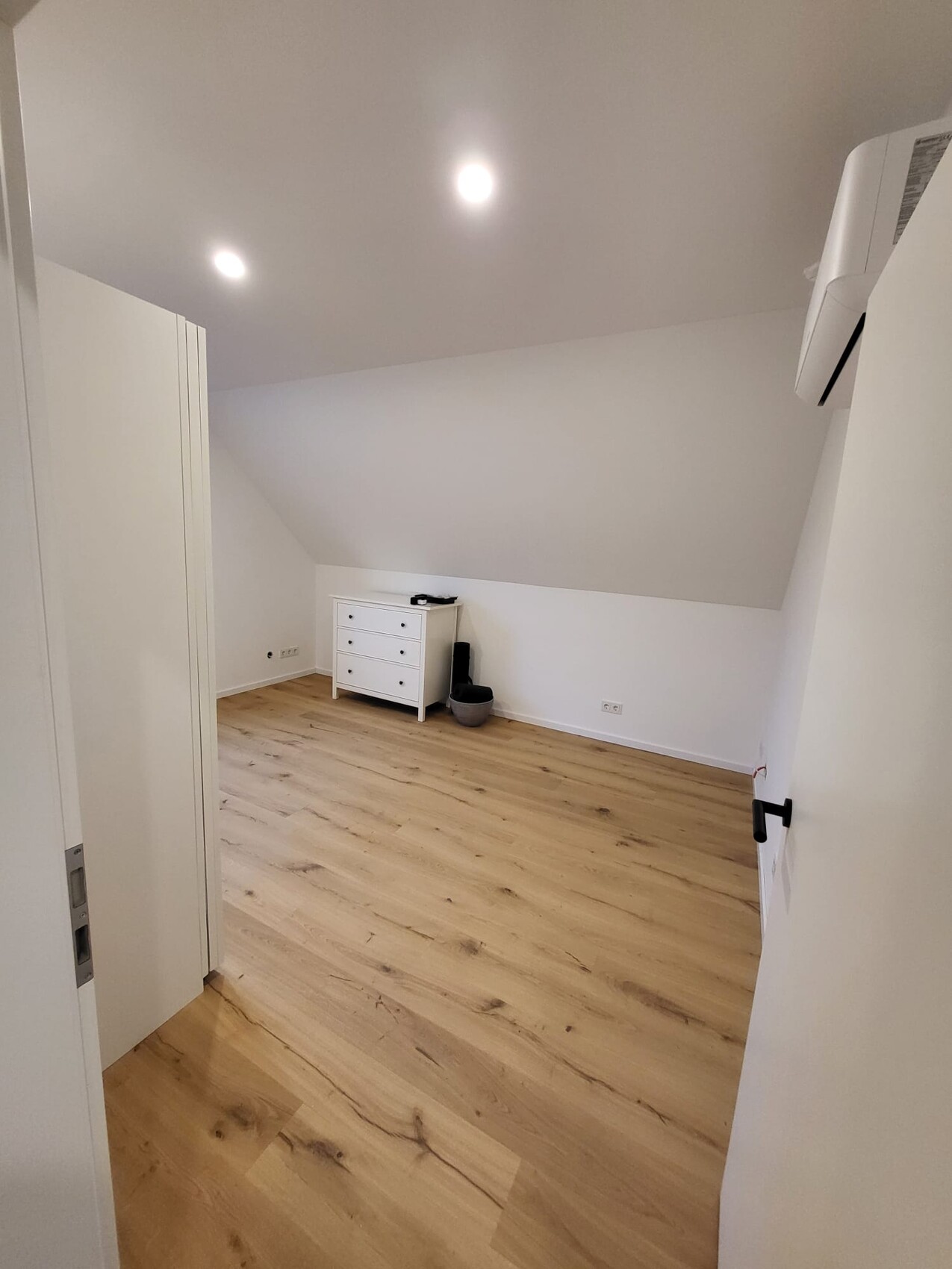 Kleiner Dachgeschoss-Wohnraum mit hellem Laminatboden, weißen Wänden, Kommode und Schrank.