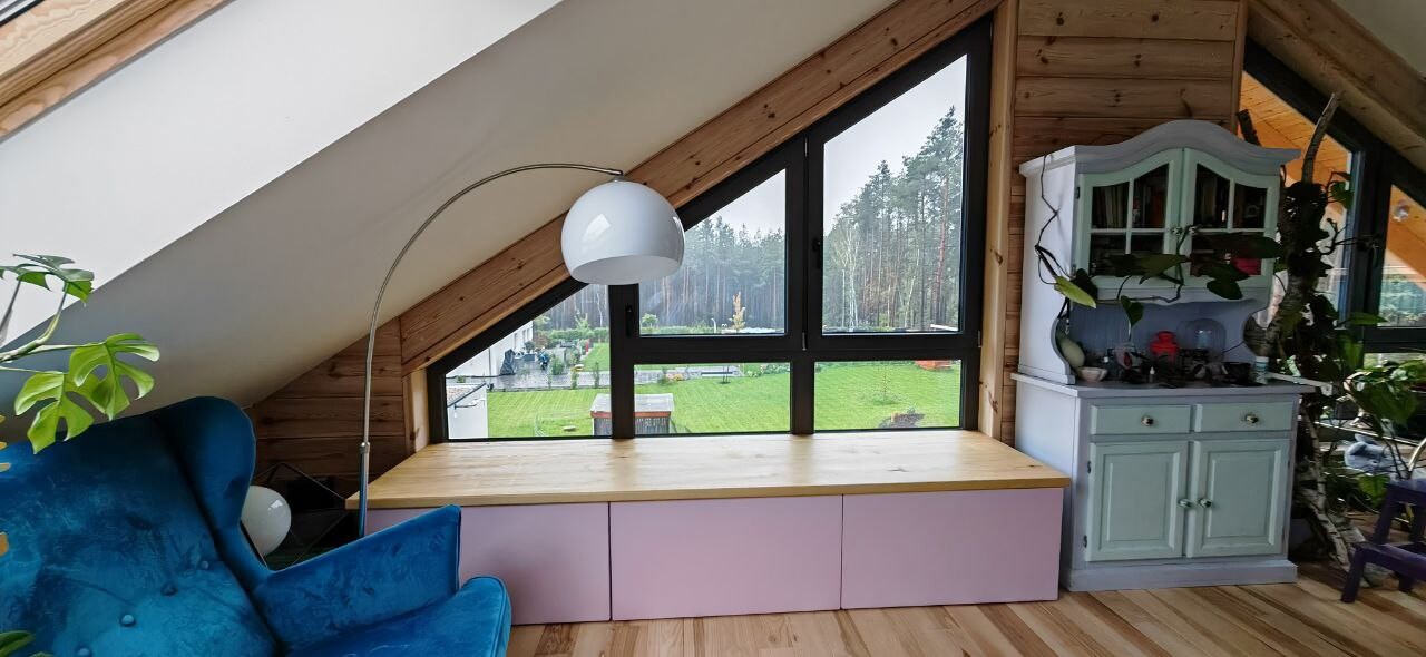 Dachgeschoss mit großem Dreiecksfenster, blauem Sessel, Pflanzenschrank und Gartenblick.