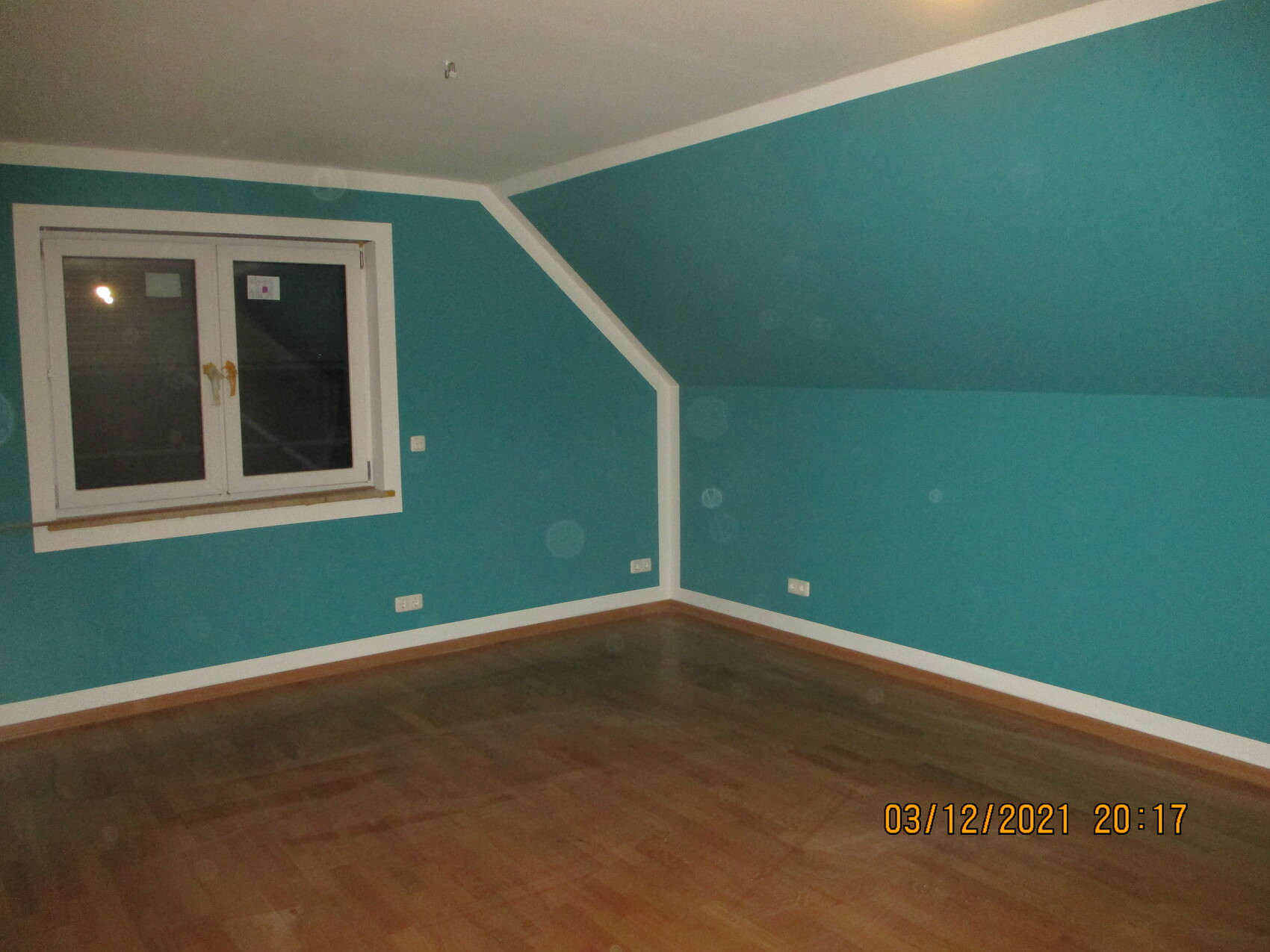 Leerer Dachbodenraum mit türkisblauen Wänden, schräger Decke, weißem Fensterrahmen und Holzboden.