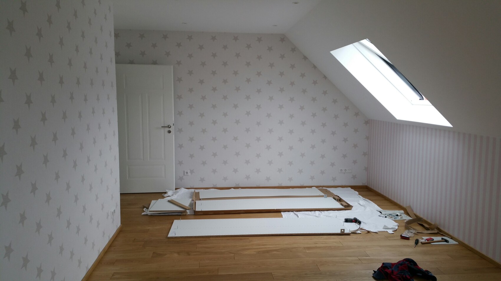 Helles Zimmer mit Sternen-Wandtapete, Dachfenster und zerlegte Möbelteile auf dem Boden.