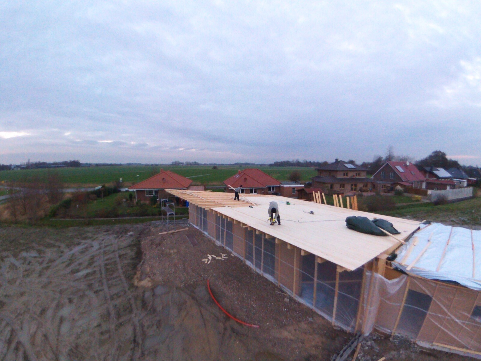 Auf einem Neubau wird eine Holz-Dachkonstruktion von Bauarbeitern errichtet, umliegende Häuser sichtbar
