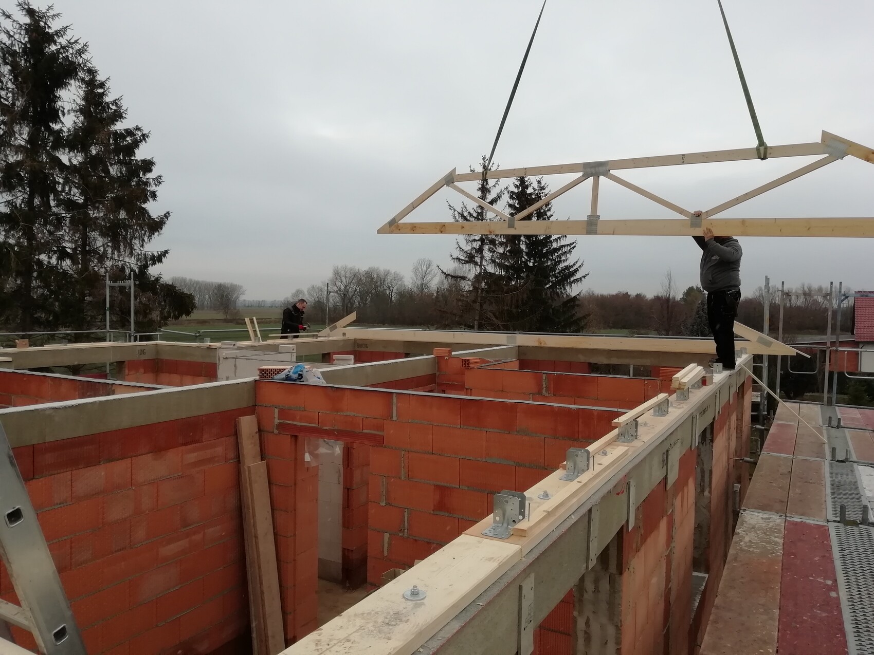 Bauarbeiter montieren Holzbalkenkonstruktion auf rotem Ziegelrohbau.
