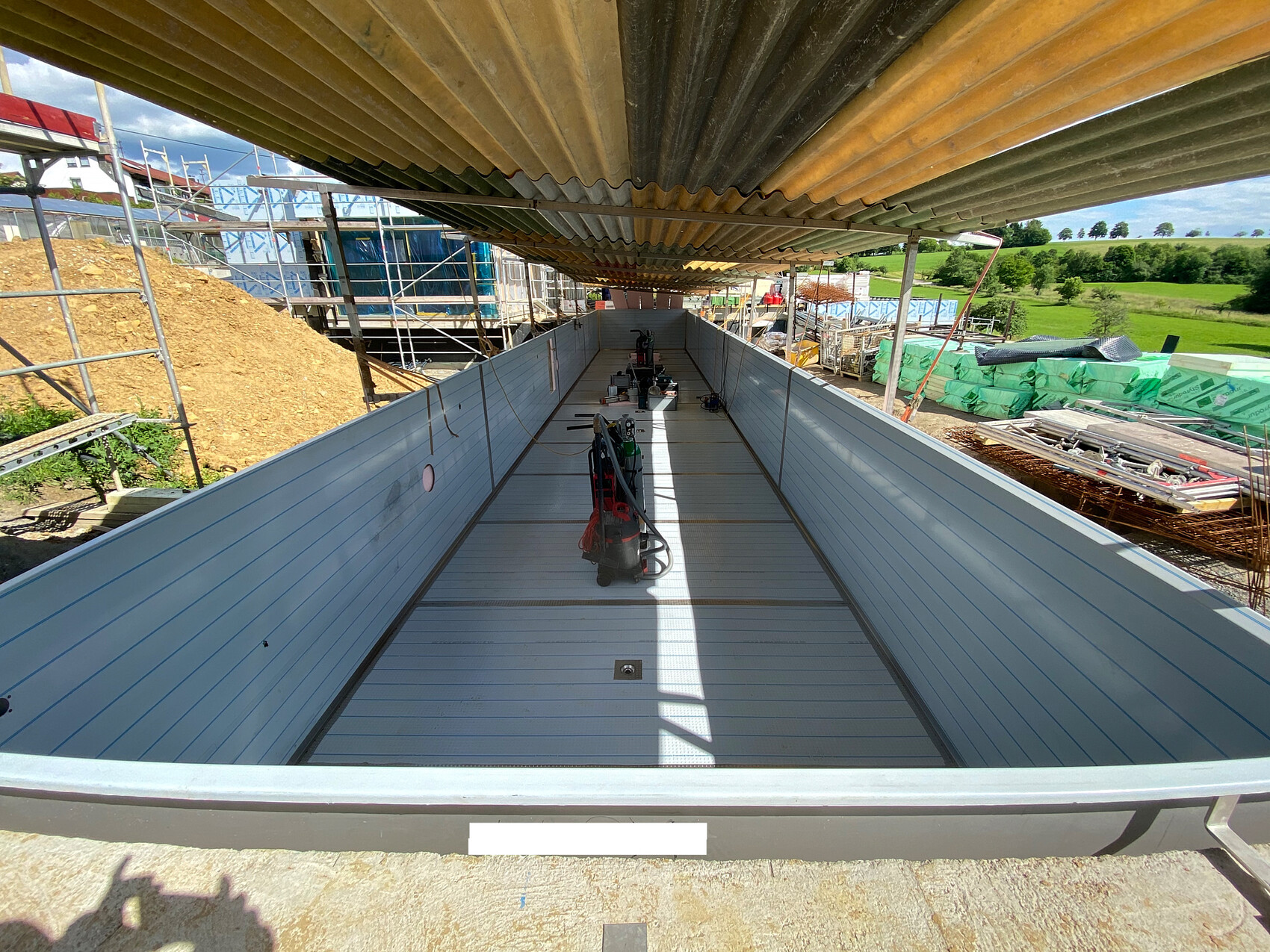 Langer offener Metallbehälter auf Baustelle mit Blechwänden, Gerüsten und Wellenblechdach.