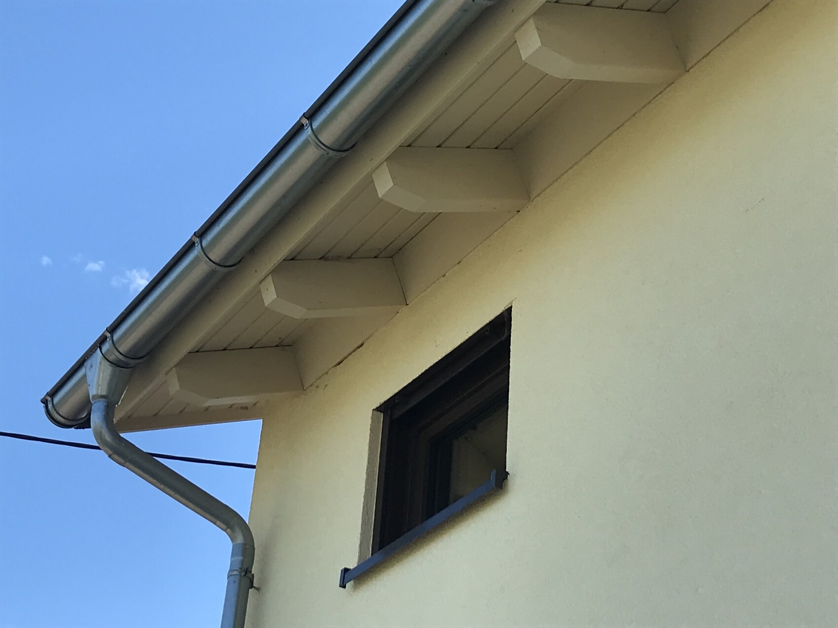 Seitenansicht eines gelben Hauses: weisse Dachrinne, Fallrohr, kleines Fenster, blauer Himmel.