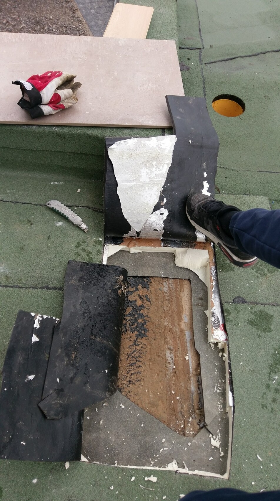 Fuß einer Person auf grünem Dachdach mit abgerissenem Material und Dämmung sichtbar.