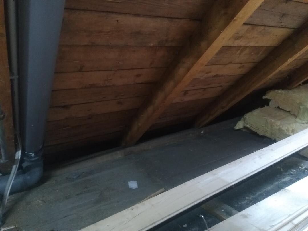 Dachboden mit Holzbalken, lose Dämmung und einem schwarzen Abflussrohr an der linken Wand.