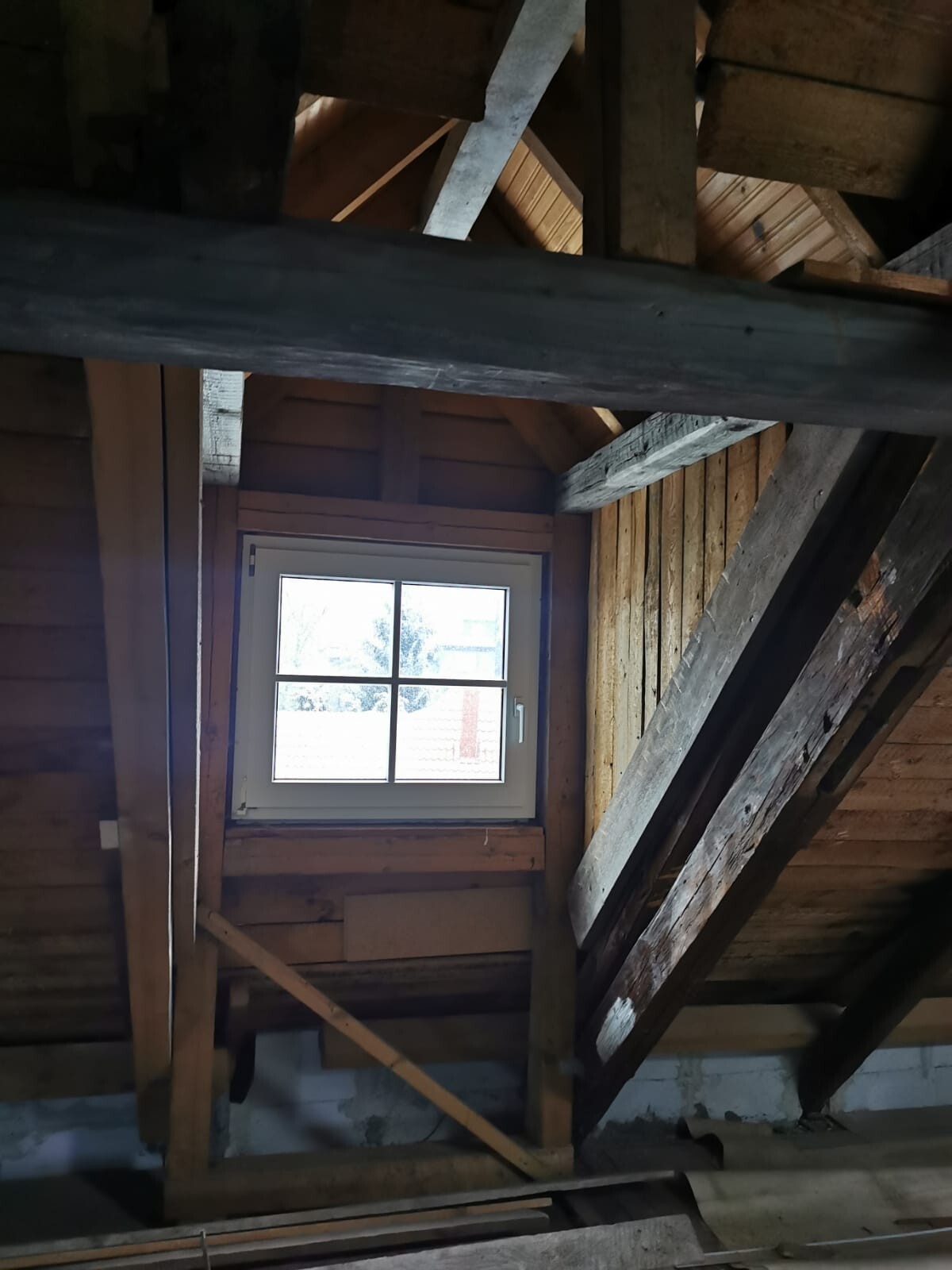 Dachboden mit sichtbaren Holzbalken und Sparren; kleines Fenster lässt Licht herein.