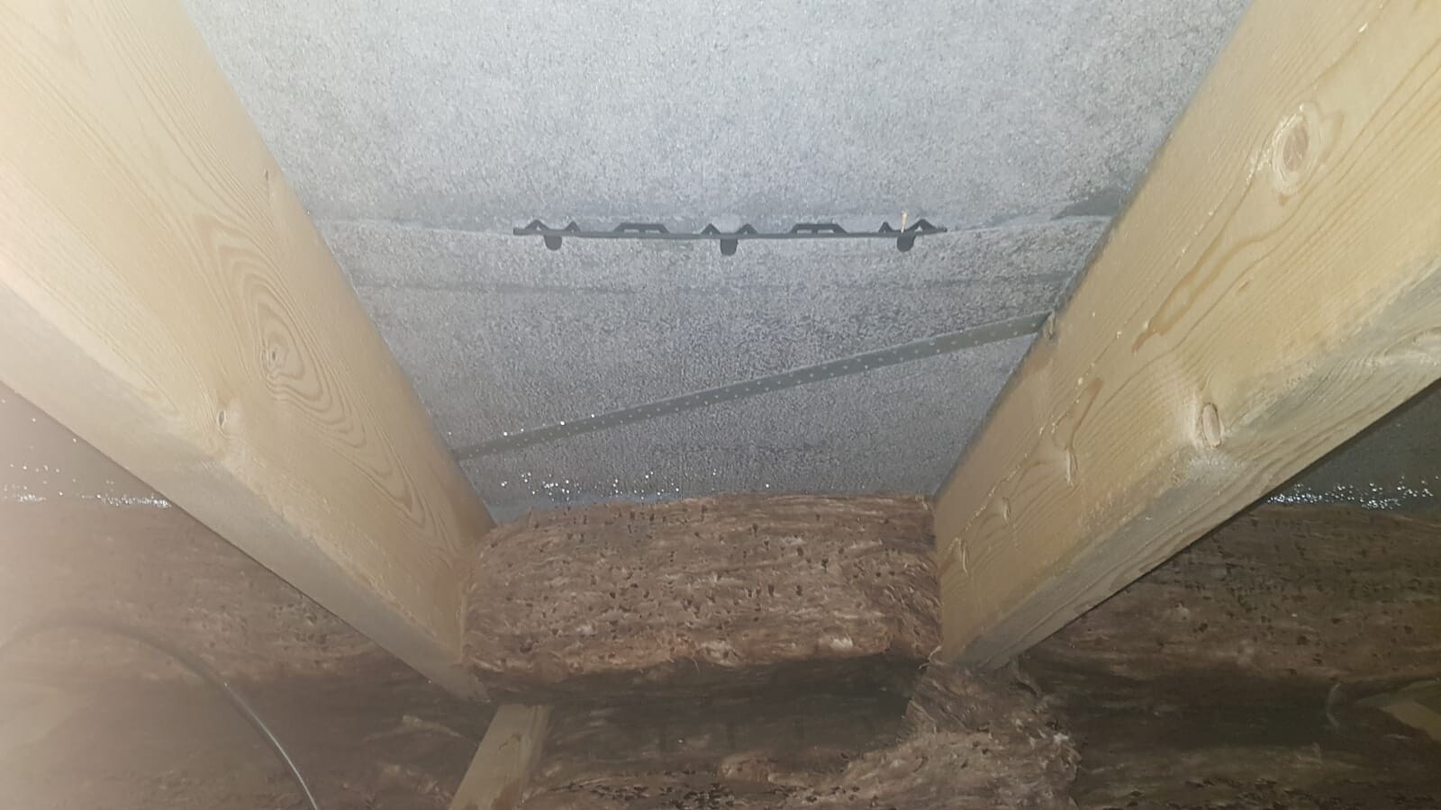 Dachbodenansicht: zwei Holzbalken, graue Deckenoberfläche, schwarze Metallstange mit Halterungen.