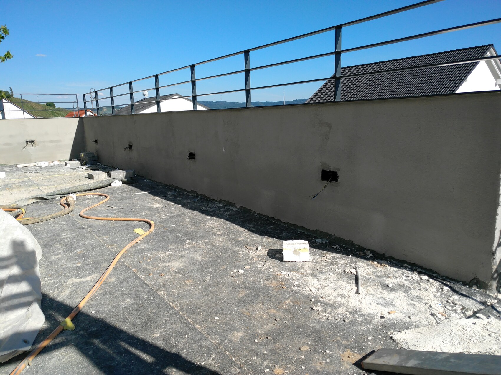 Dachterrasse im Bau: Betonwand mit Kabelauslaessen, Schlauch und Baustellenabfall, blauer Himmel.