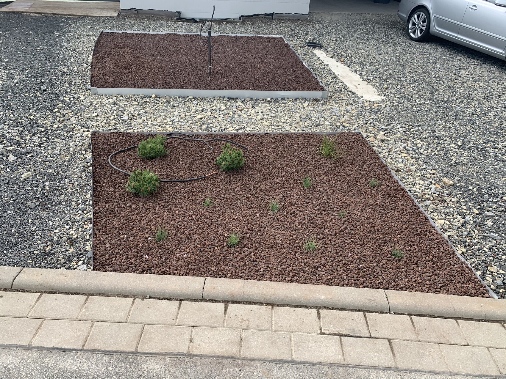 Vorgarten mit rotem Kies, kleinen Grasbüscheln, Gartenschlauch, geparktem Auto.