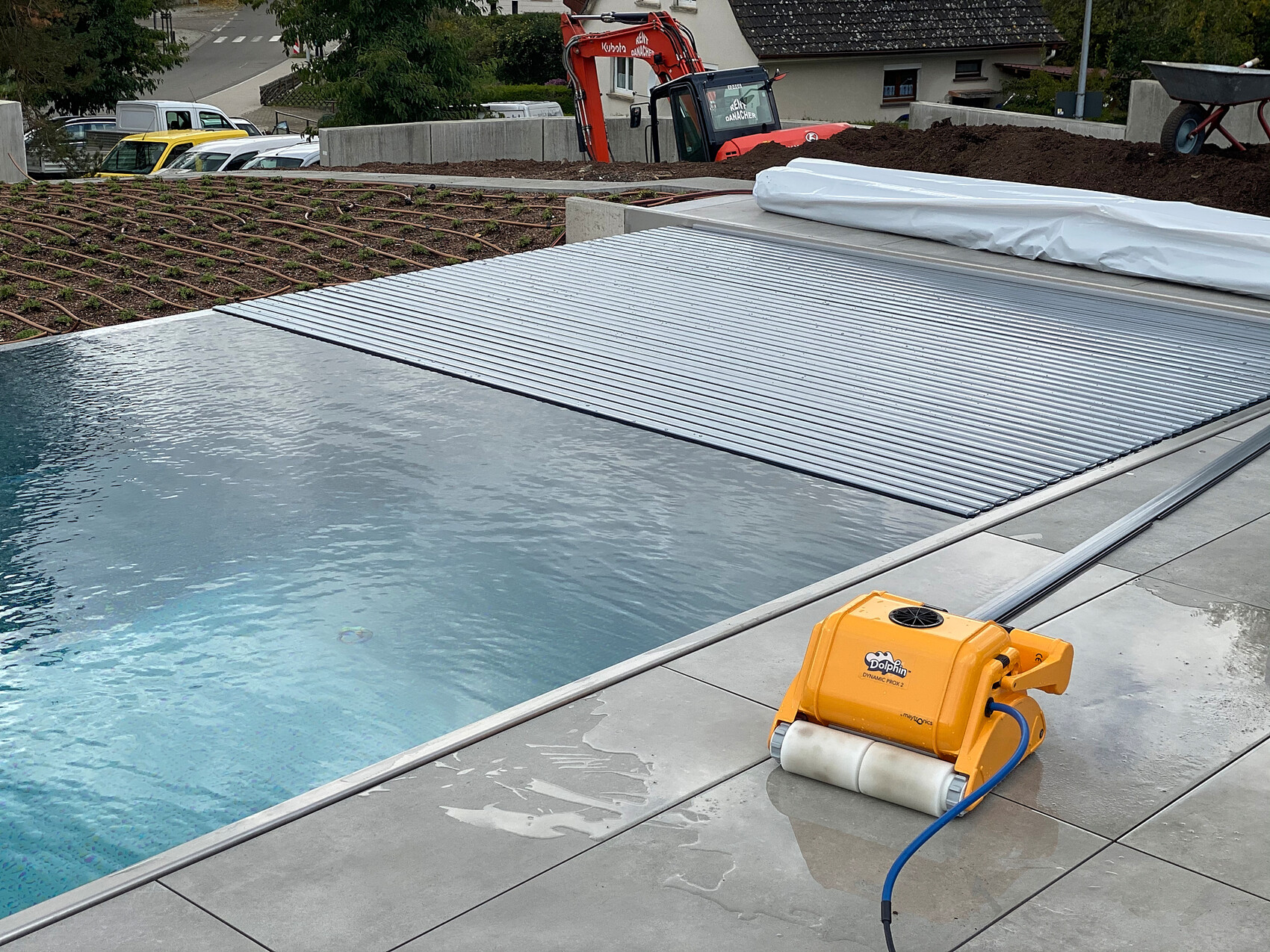 Bauarbeiten an einem Pool: graue Platten, gelbe Poolabdeckrolle, Abdeckung bereit, Bagger.