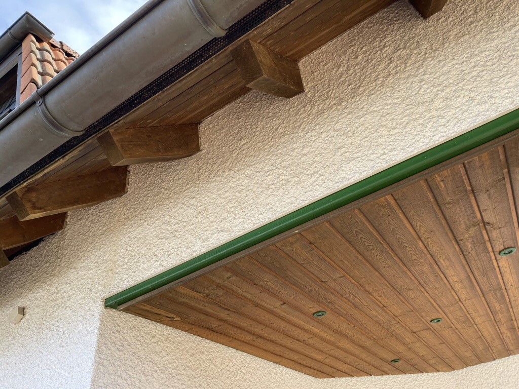 Unter dem Dachüberstand sichtbare Holzbalken, Dachrinne, Putzwand und eine Holzdecke.
