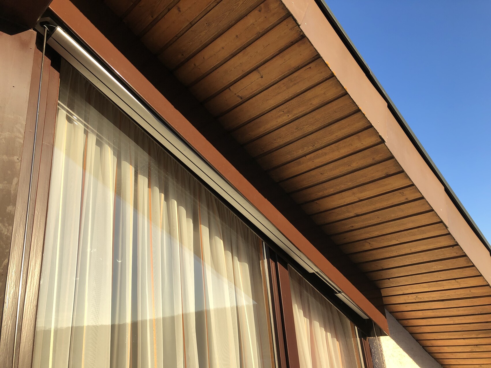 Außenansicht eines Holzhauses mit Fenster und Vorhängen, überstehendes Holzdach, blauer Himmel.