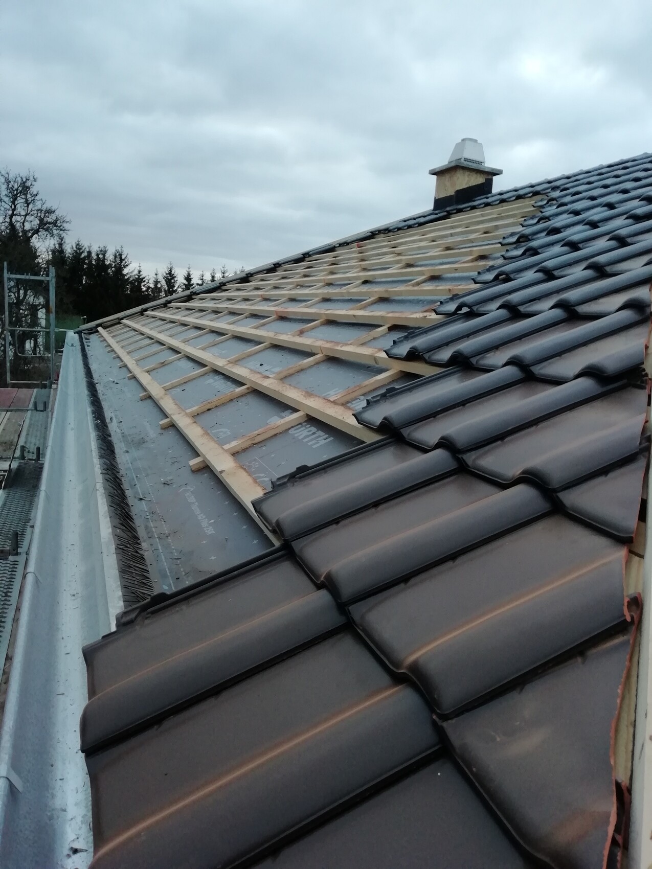 Teilweise gedecktes Dach: Holzlattenunterkonstruktion, dunkle Dachziegel, Kamin, grauer Himmel.