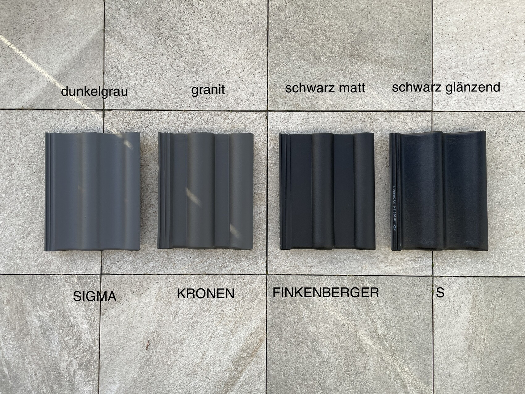 Vier Dachziegel-Proben in Dunkelgrau, Granit, Schwarz matt, Schwarz glänzend auf Steinboden.