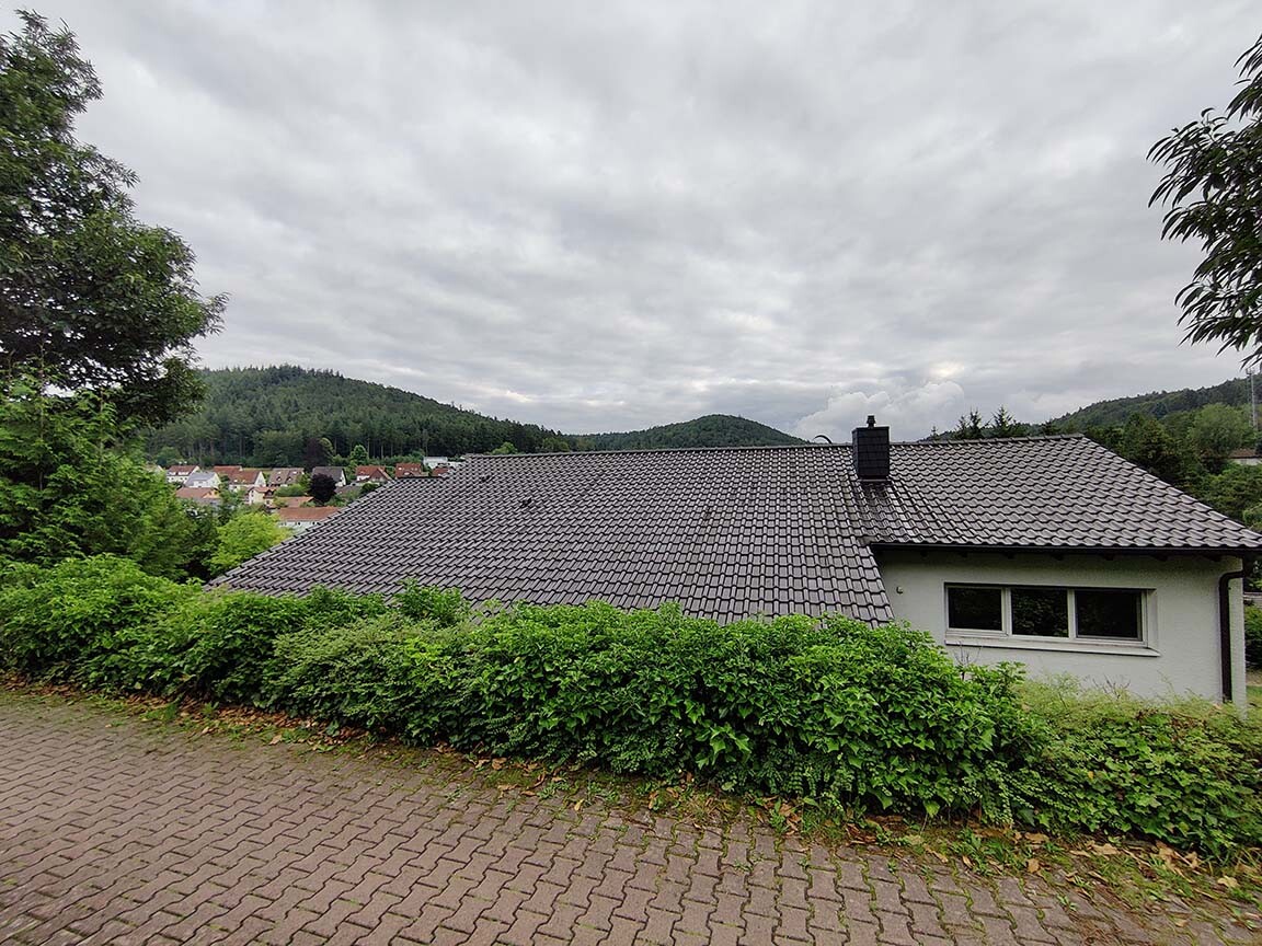 Häuschen mit grauem Ziegeldach, grüne Hecke davor, gepflasterter Weg, Waldhügel im Hintergrund.