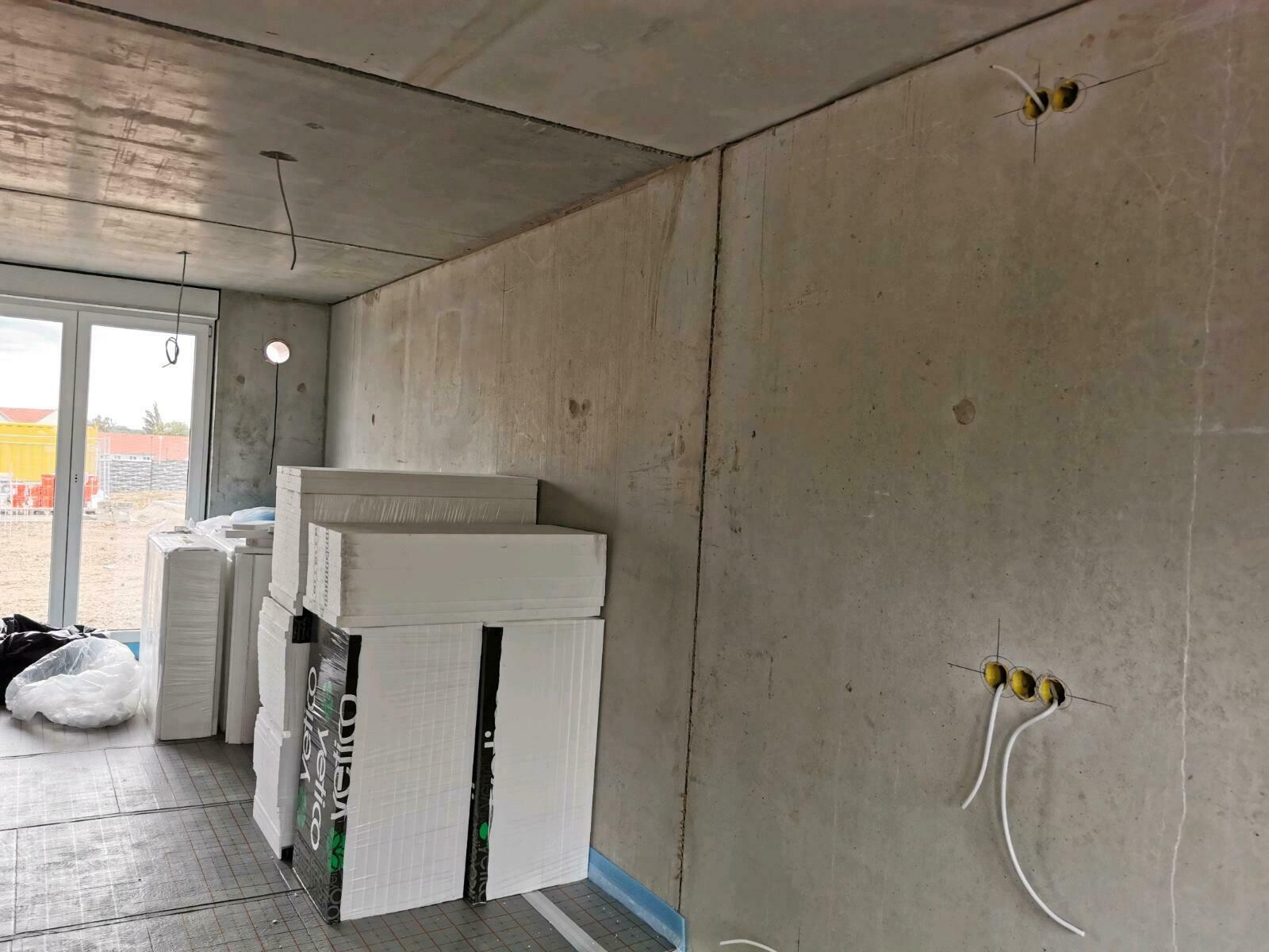 Rohbauzimmer: Betonwände, Fensterfront, gestapelte Dämmplatten und freiliegende Kabel.