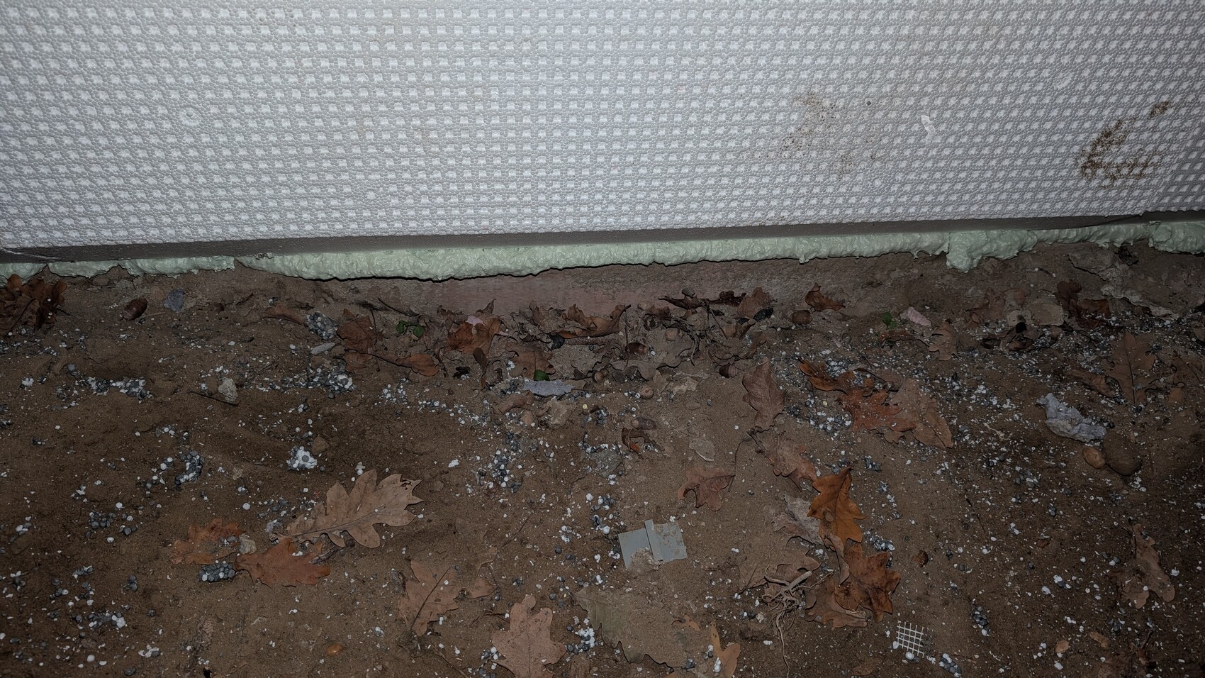 Außenbereich: Boden mit Kies und Laub, sichtbare Dämmung am Fundament der Wand
