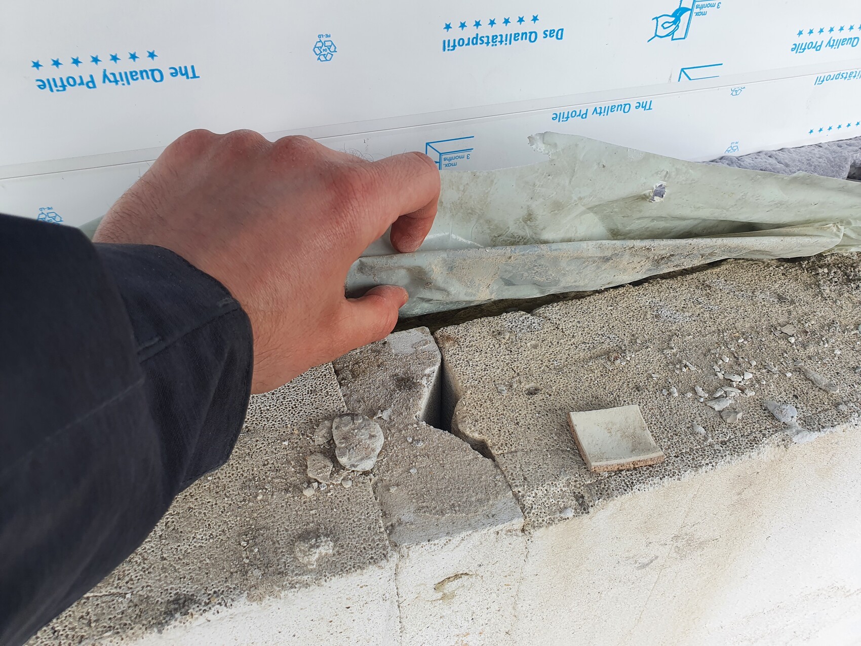 Nahaufnahme einer Hand, die Baufolie an einer Betonmauer-Fuge festhält; daneben Kies und Bruchstücke.