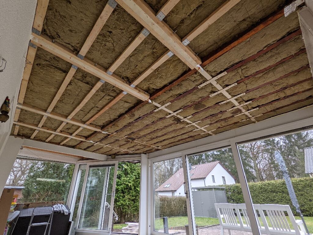 Offene Deckenstruktur mit Dämmung im Wintergarten; Blick auf Garten und Nachbarhäuser.