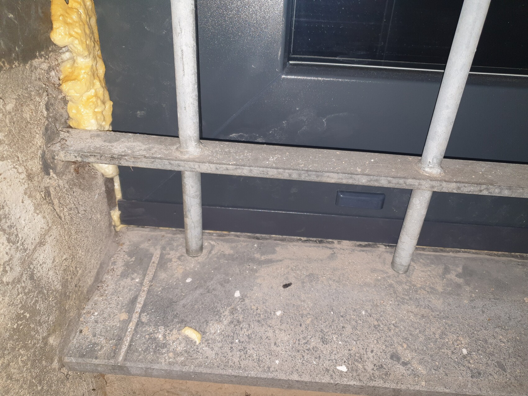 Dunkle Tür hinter Metallgeländer mit Stäben; gelber Isolationsschaum quillt aus Wandfuge links.
