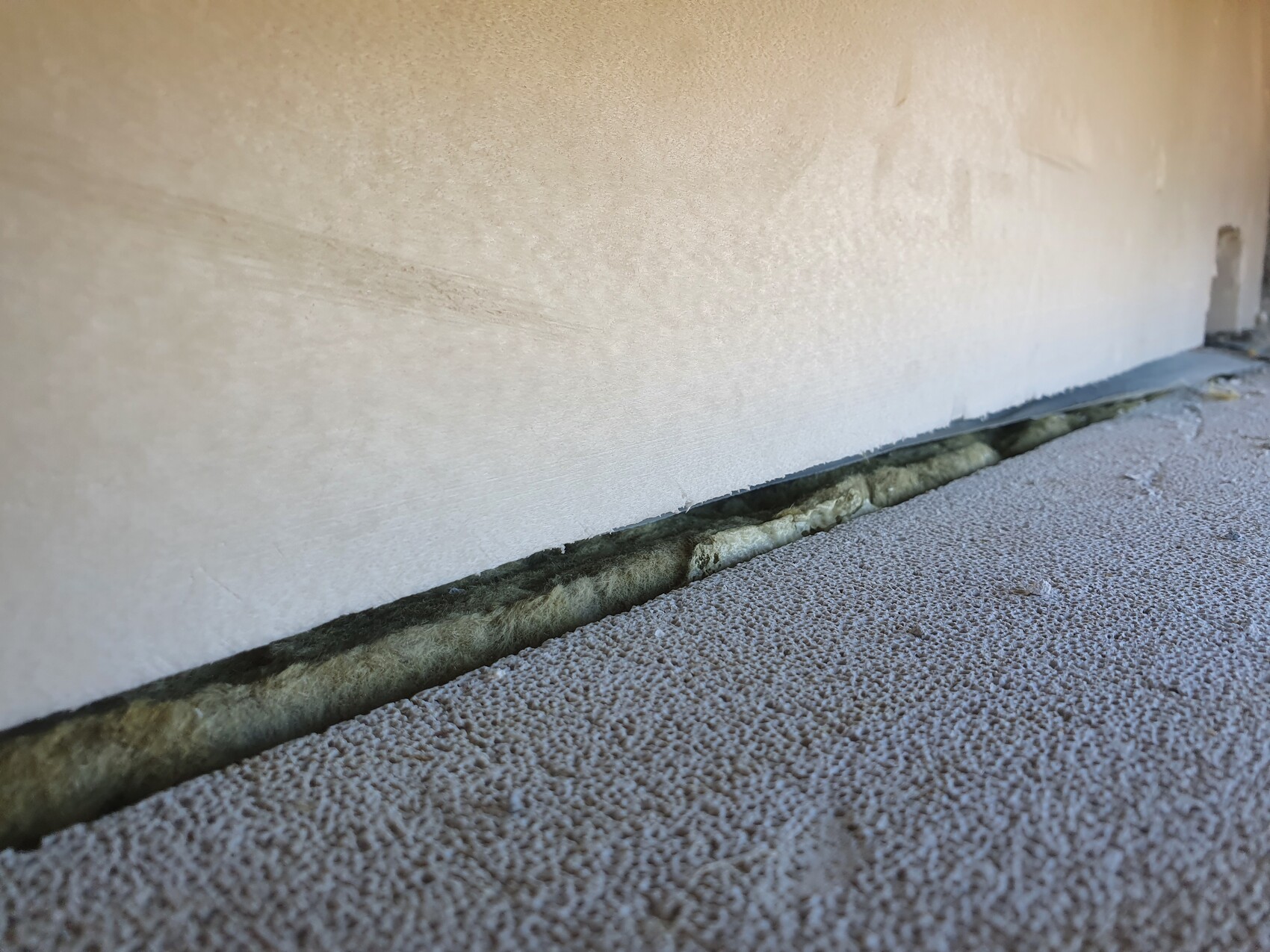 Unterkante einer hellen Wand mit kleinem Spalt zur Bodenplatte und grüner Dämmung im Spalt.