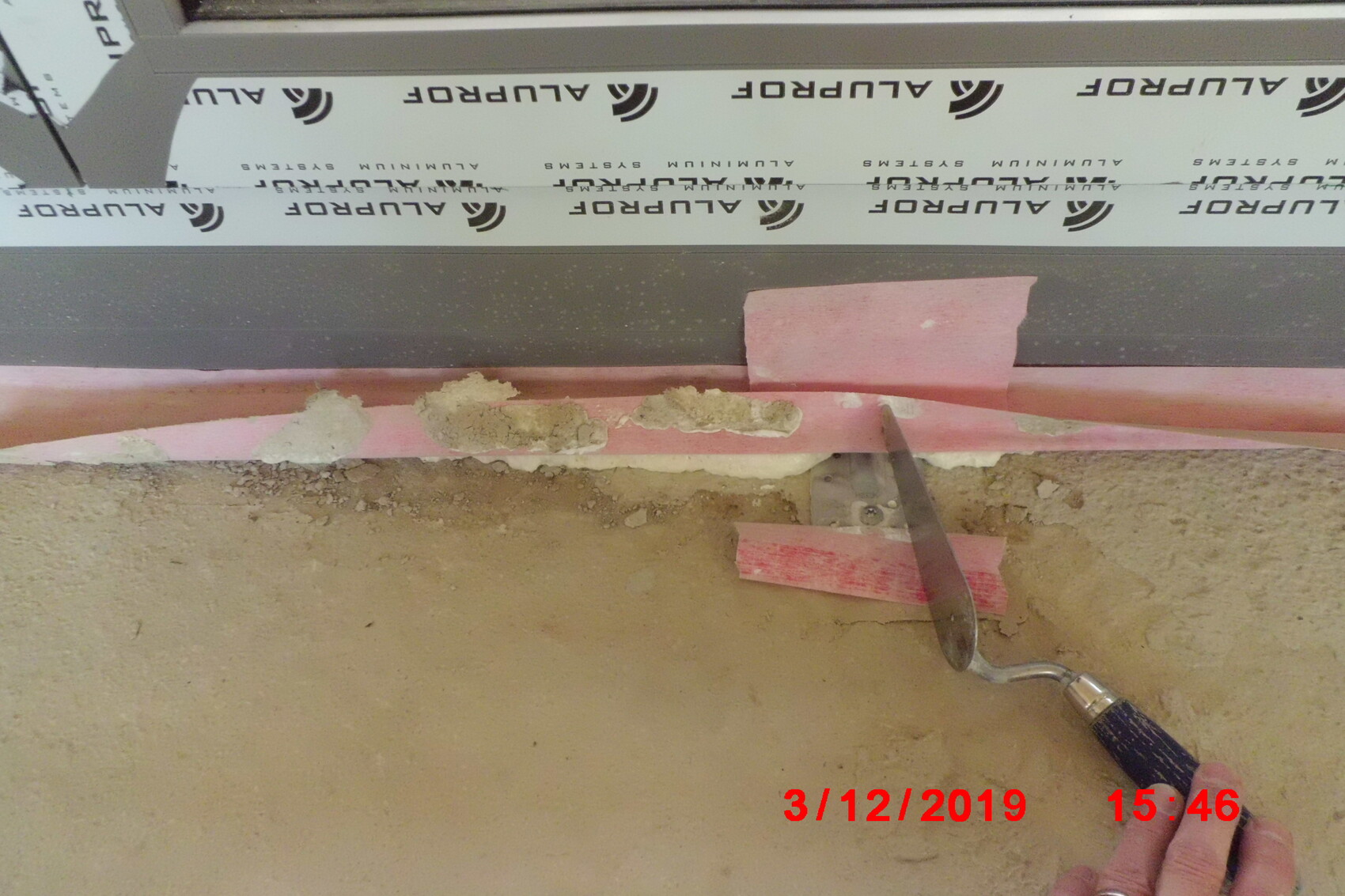 Werkzeug schneidet rosa Dämmplatten an einer Wandkante bei Bauarbeiten, Erde und Staub sichtbar.