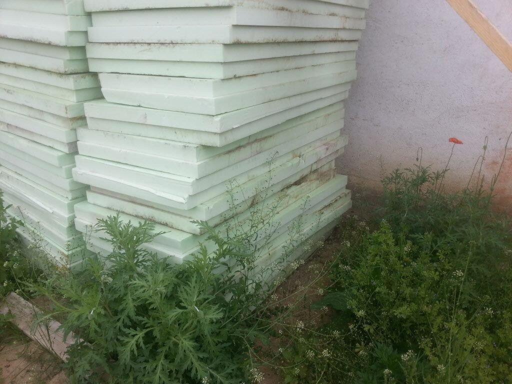 Stapel grünlich-weißer Isolierplatten aus Styrofoam im Freien neben einer Wand, von Unkraut umgeben.