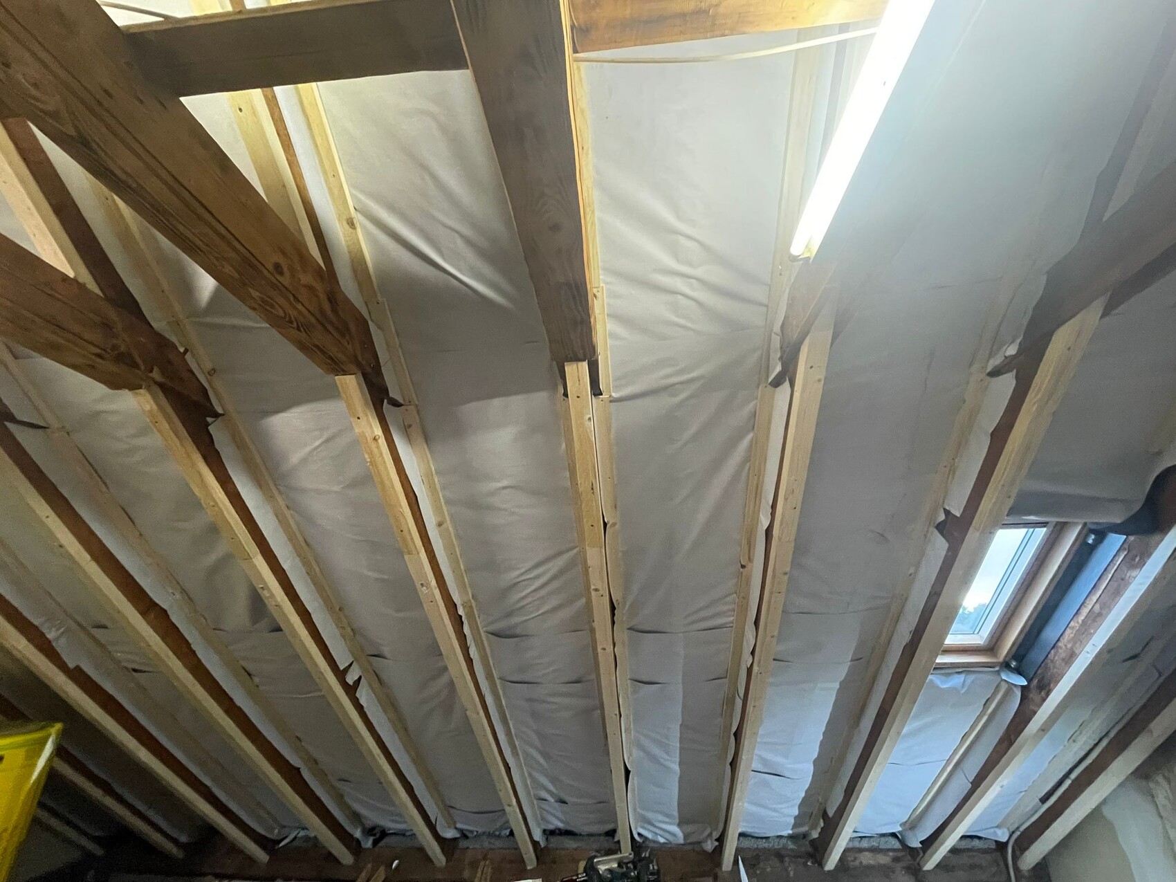 Dachboden mit freigelegten Holzbalken, Dämmung dazwischen, Fenster rechts und Leuchtstofflampe.