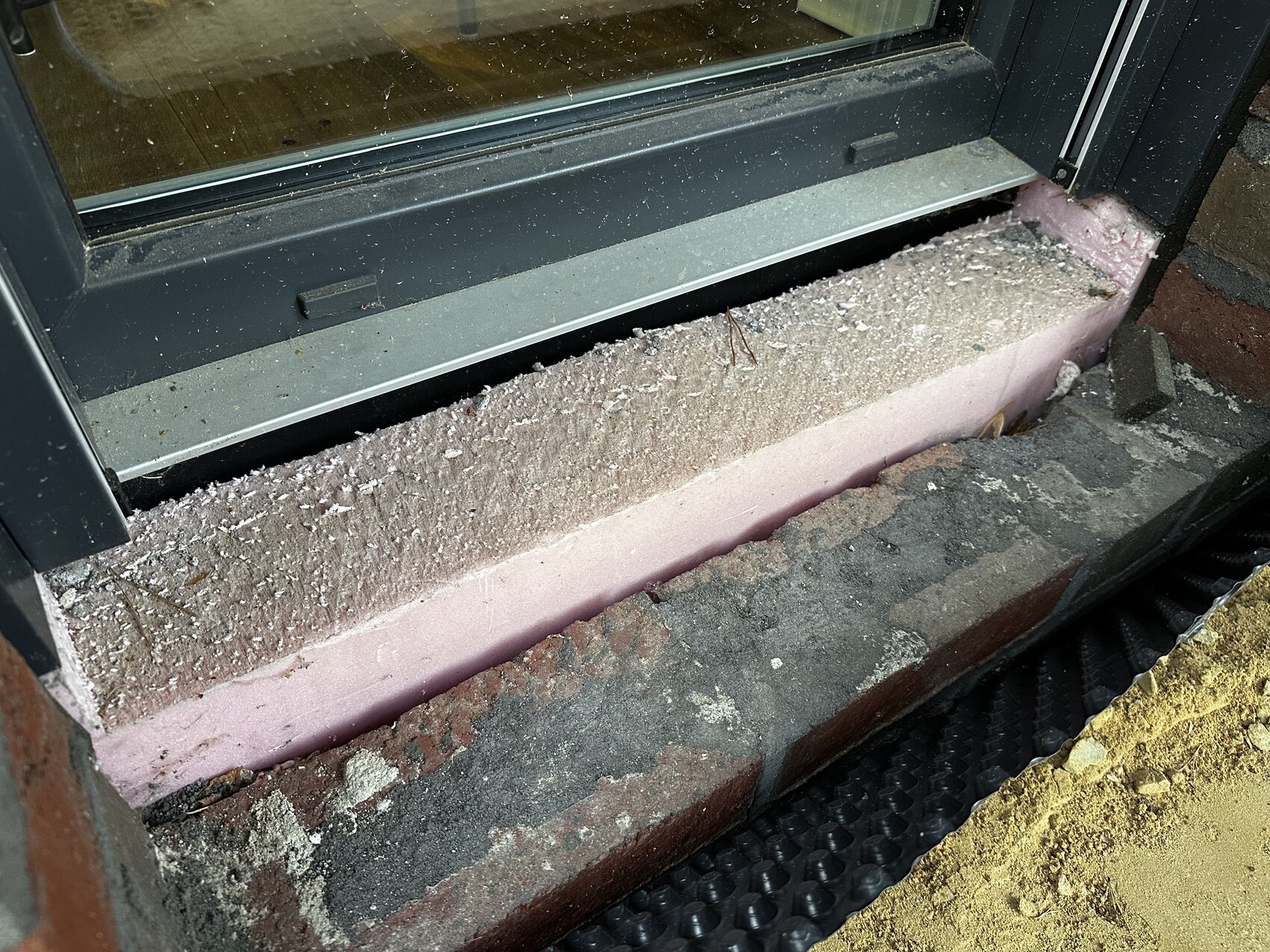 Unterseite eines Fensterrahmens mit rosa Dämmplatte, Staub und Schutt am Boden.