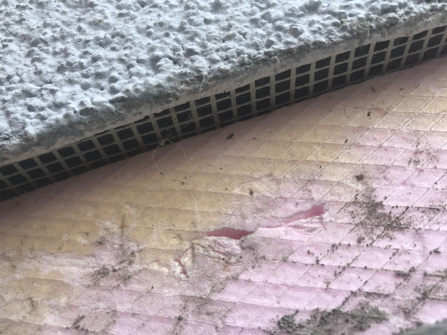 Nahaufnahme eines Lüftungsgitters unter einer groben Betonkante über pink-beigen Fliesen mit Schmutz.