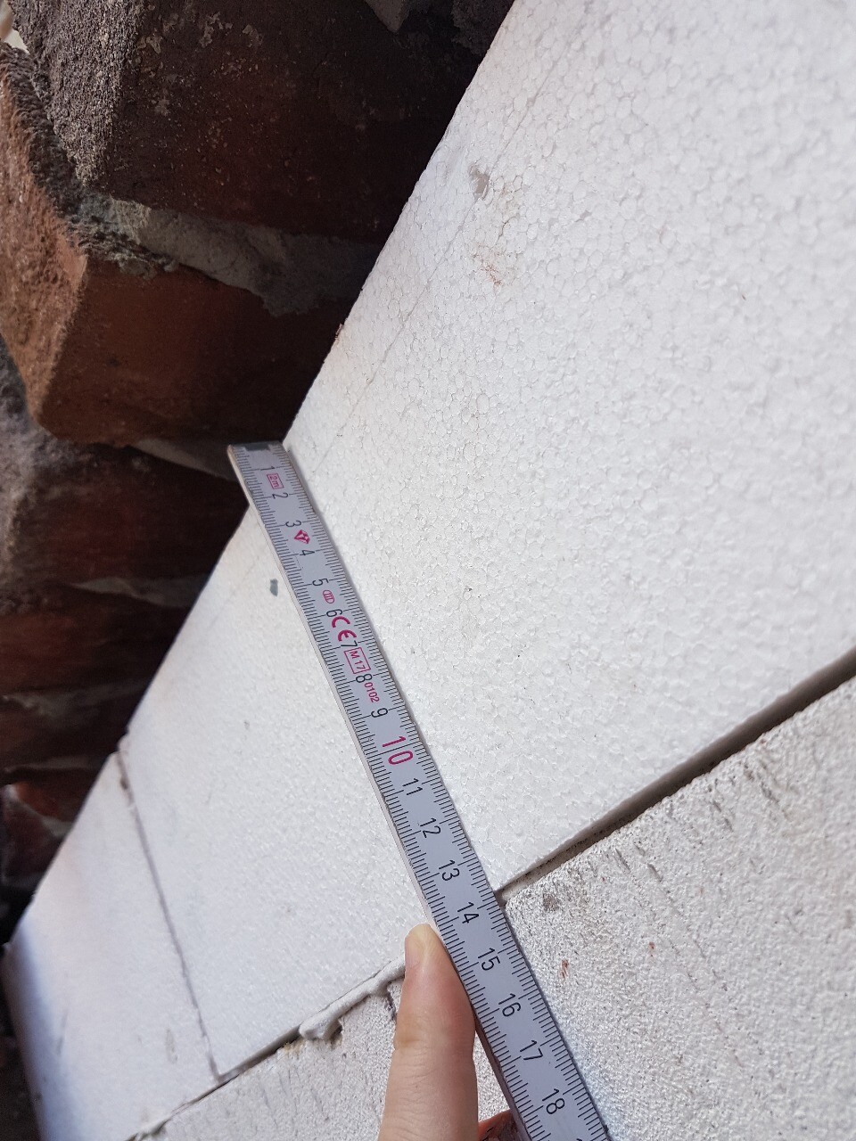 Hand hält Maßband an weisse Dämmstoffwand, roter Backstein im Hintergrund