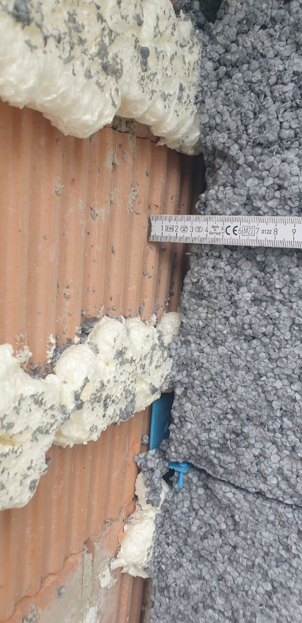 Nahaufnahme einer Ziegelwand mit weißem Dämmmaterial, grauem Kies und Maßband.