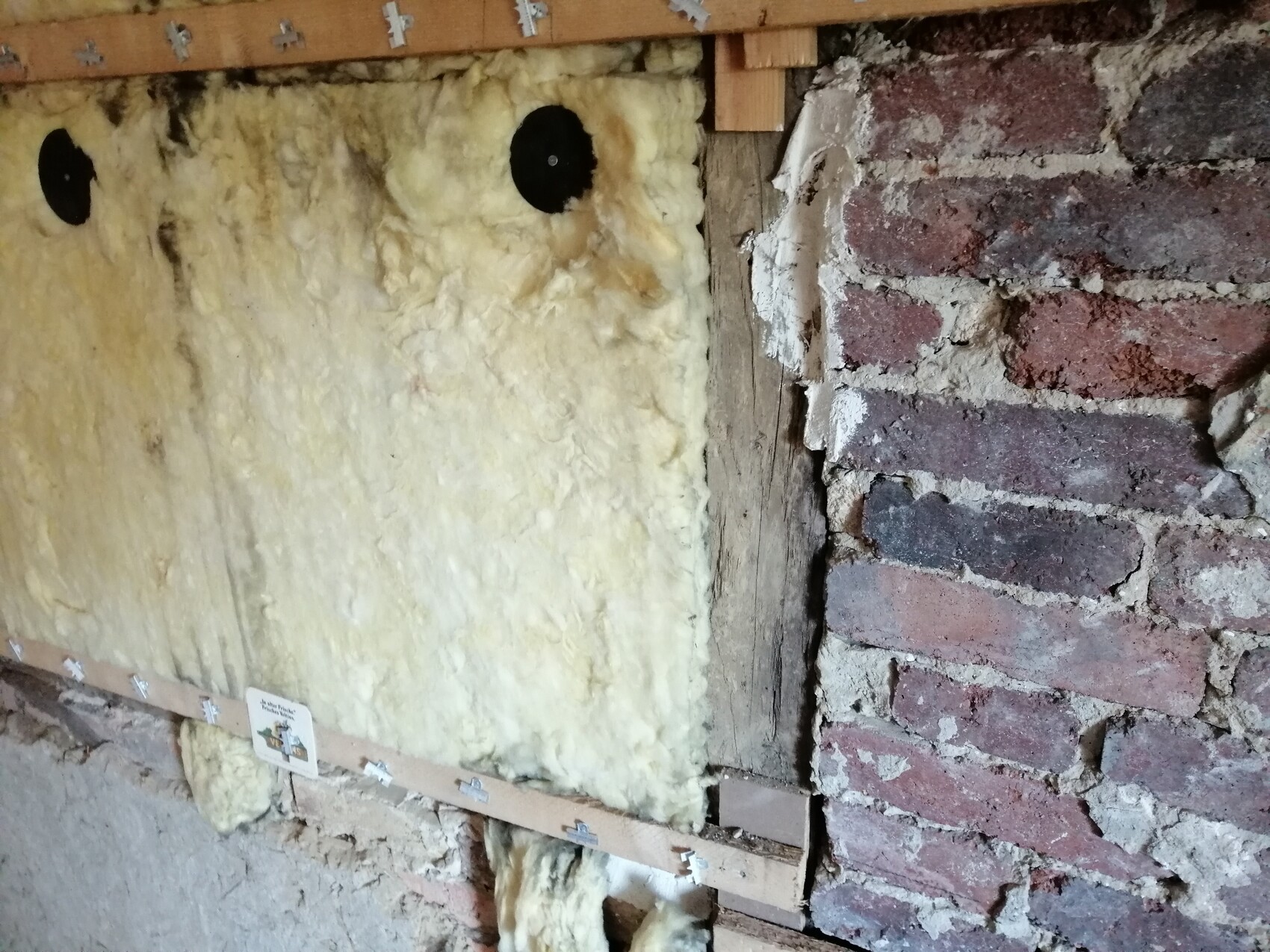 Wand mit Sprühdämmung (gelb) im Hohlraum zwischen Holzrahmen; Backsteinwand rechts.