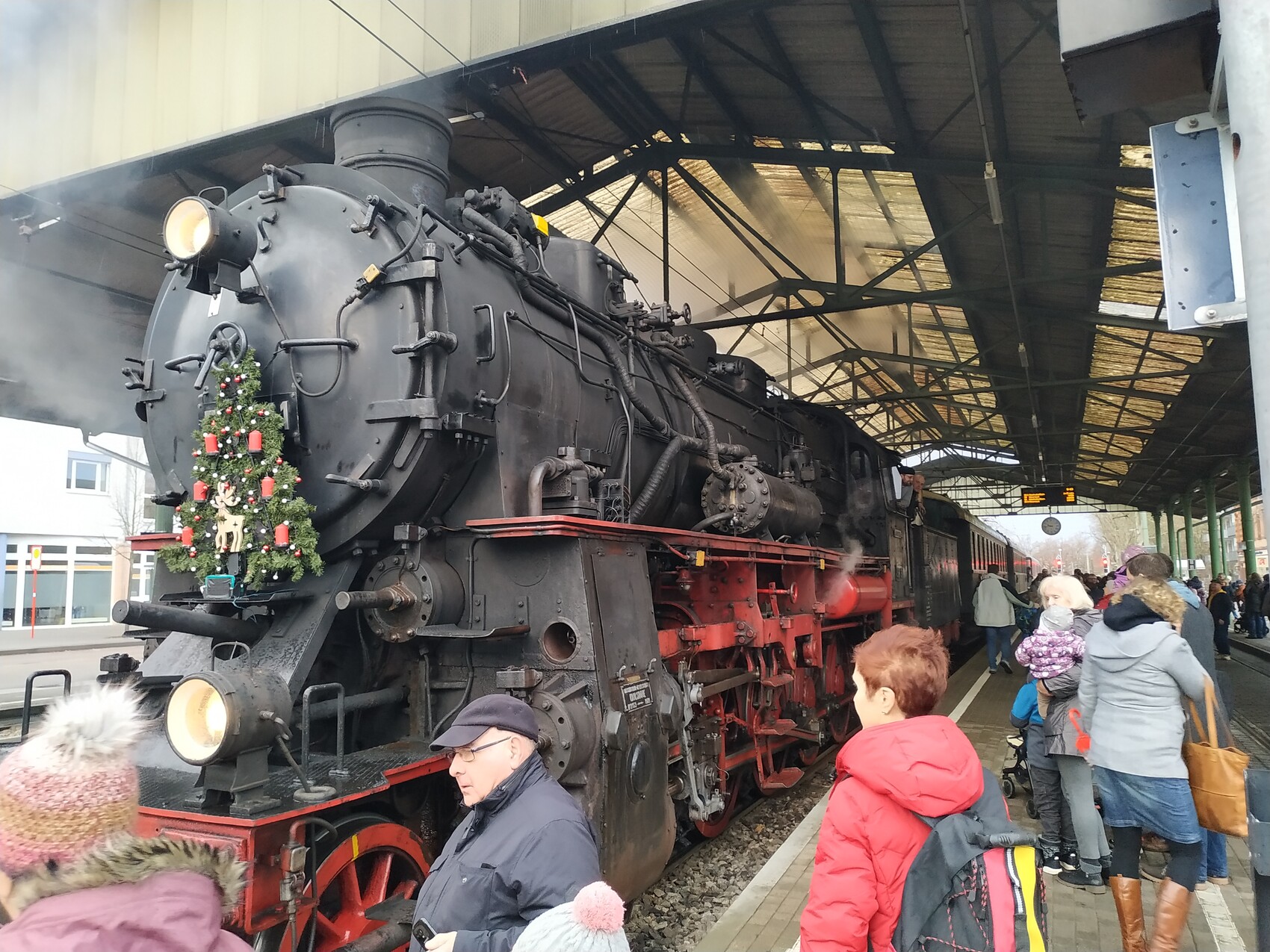Historische Dampflokomotive auf dem Bahnsteig; geschmückter Weihnachtsbaum vorne, Menschen schauen zu.