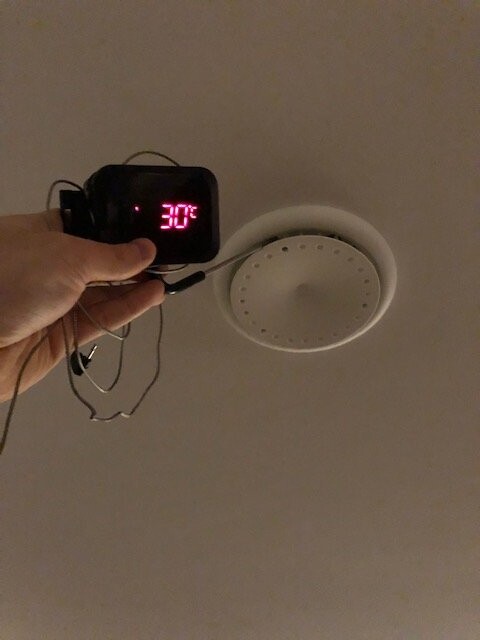 Hand hält ein digitales Thermometer mit roter Anzeige (30°) vor einem Deckenrauchmelder.