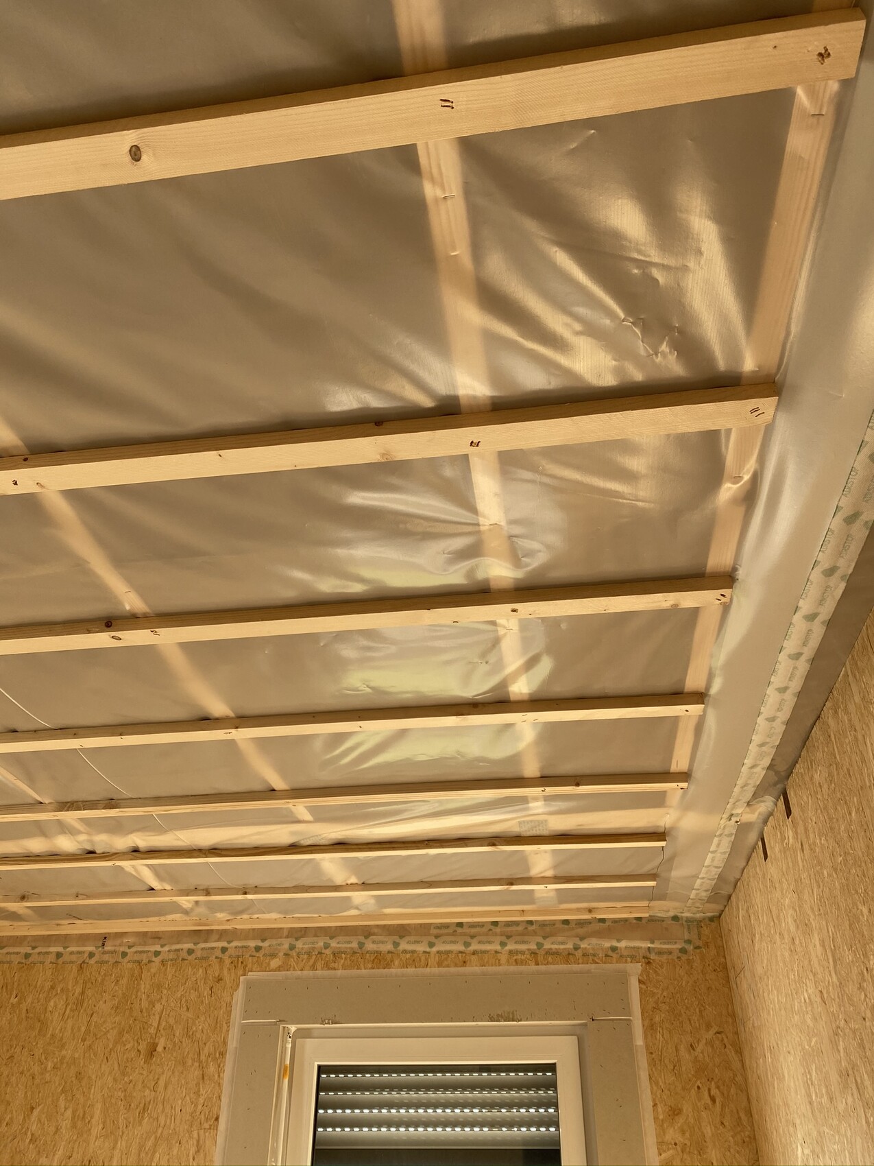 Offene Decke mit Holzlatten, Folien-Isolierung und Fenster im unteren Bereich.