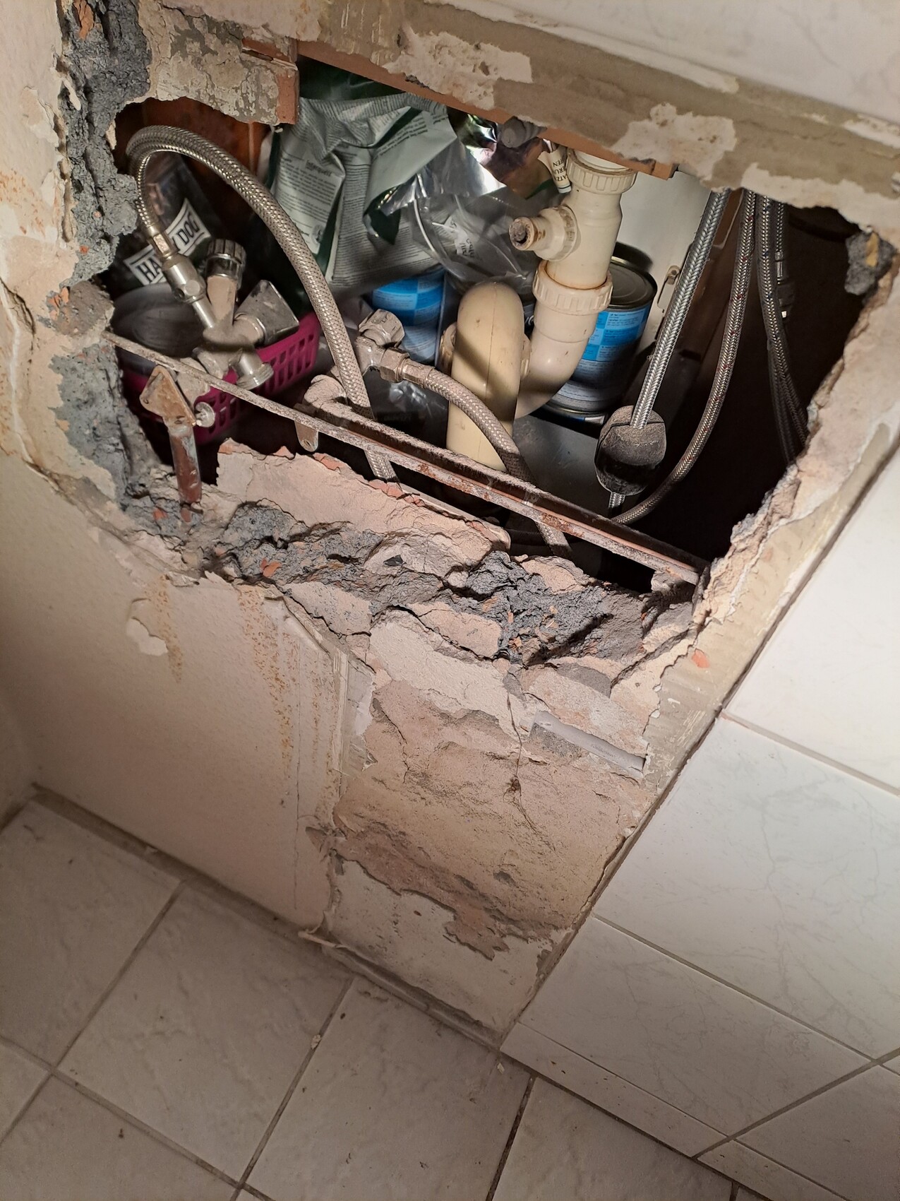 Beschädigte Wand offenbart Rohre, Schläuche und Abfluss unter dem Waschbecken.