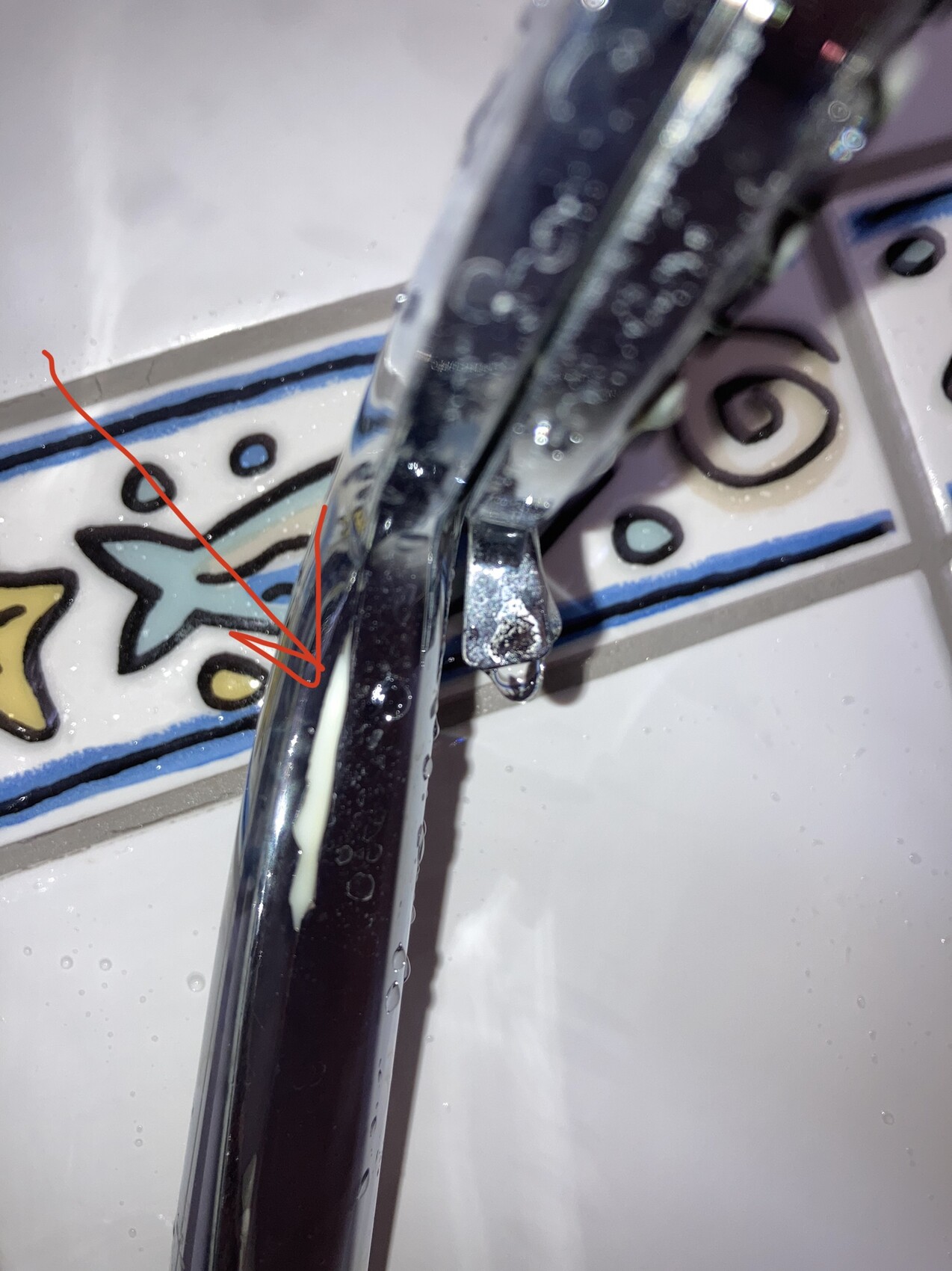 Nahaufnahme eines verchromten Wasserhahns mit Tropfen, bunte Fliesen im Hintergrund.
