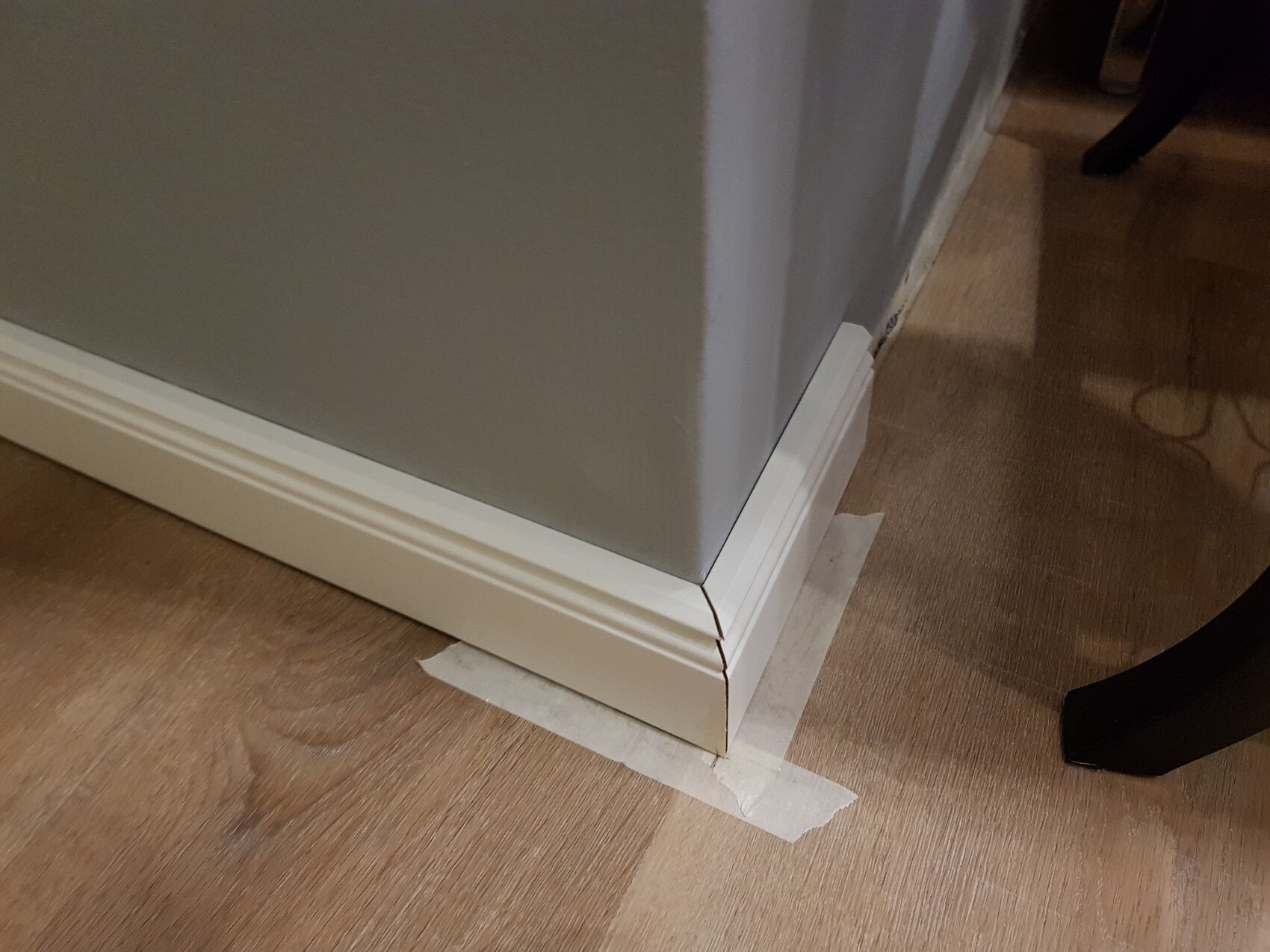 Ecke einer grauen Wand mit weißer Sockelleiste; Riss in der Leiste und Malerband am Boden.