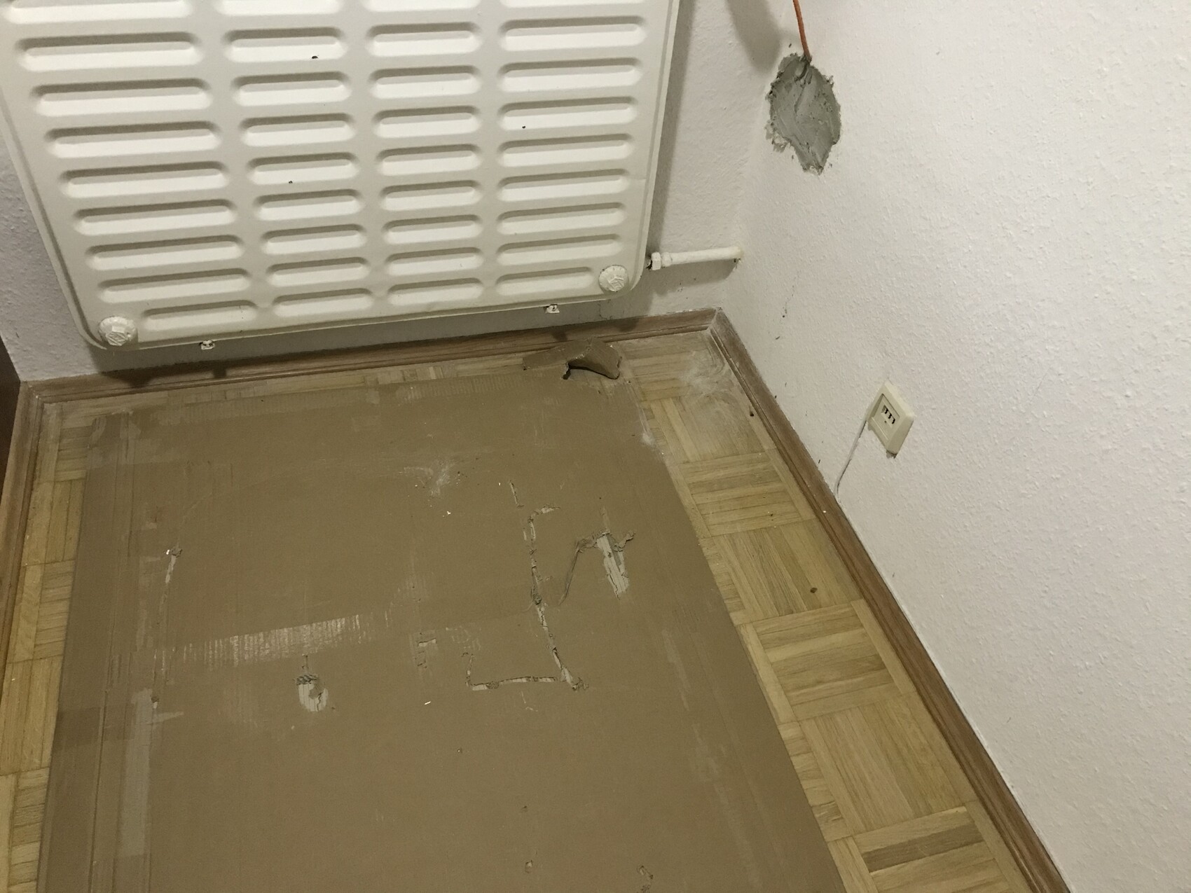 Heizkörper an der Wand; Boden mit brauner Platte, beschädigte Wand mit offener Kabelöffnung.