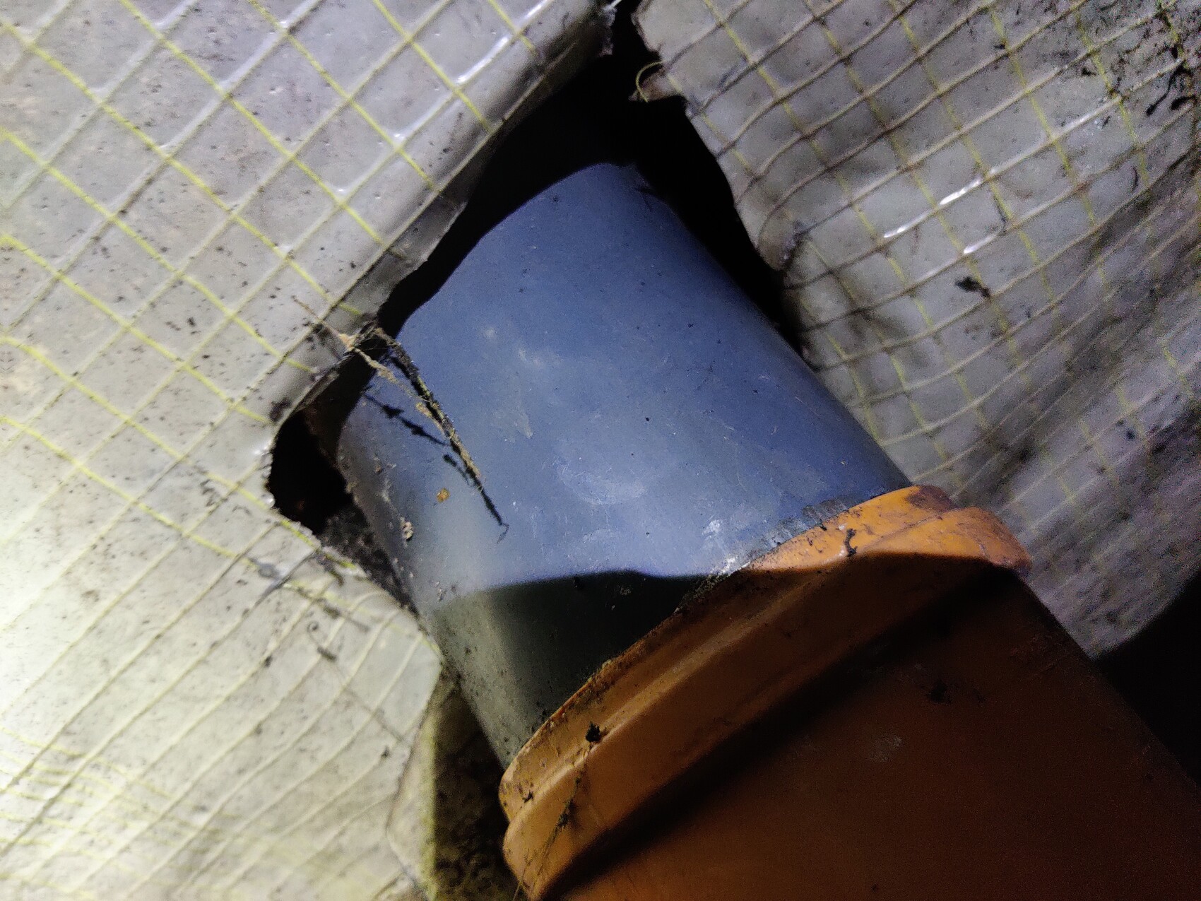 Offenes Loch in Betonwand, durch das ein blaues Rohr sichtbar ist; Rand mit orangefarbener Schelle.