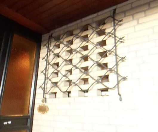 Metallgitter mit diamantförmigen Öffnungen an weißer Ziegelwand; links Fenster mit orangeglas.