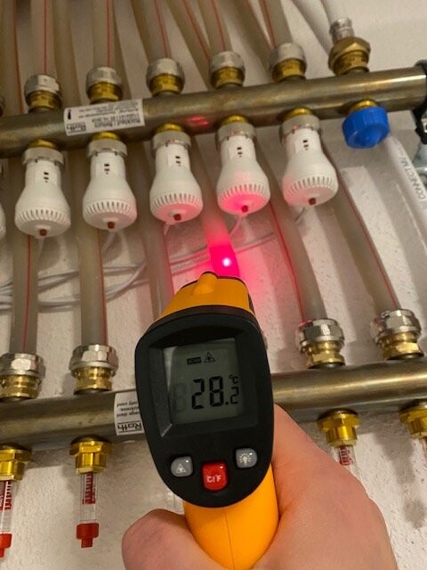 Hand hält Infrarot-Thermometer an Heizungsrohren; Anzeige 28,2°C, roter Lichtpunkt.