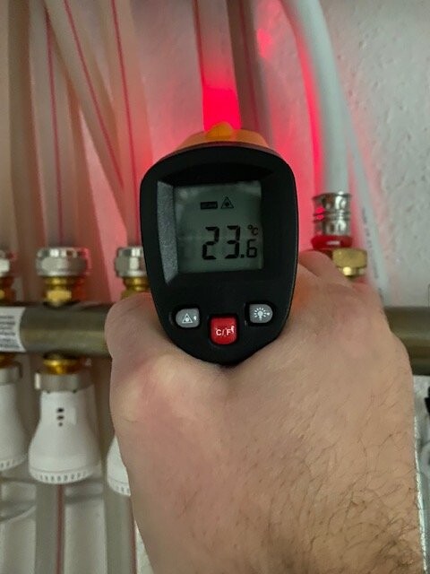 Hand mit digitalen Temperaturmessgerät zeigt 23,6 °C, rotes Licht hinter Rohren und Ventilen.