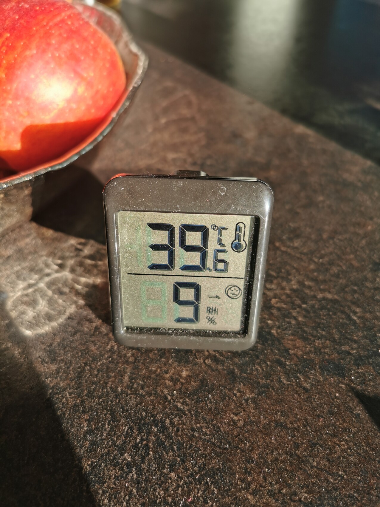 Kleines digitales Thermo-Hygro-Messgeraet auf dunkler Küchenarbeitsplatte neben einem Apfel