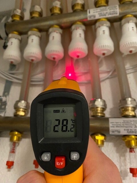 Hand mit Infrarot-Thermometer vor Heizungsrohren und Ventilen; Display zeigt 28.2°C.